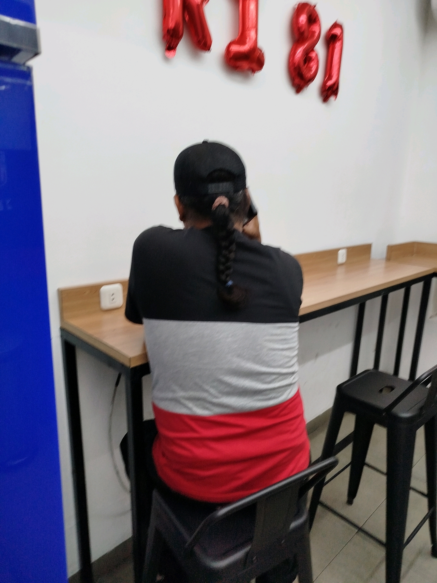 The image depicts the back of a person wearing a black baseball cap and a t-shirt with horizontal stripes of black, gray, and red. The person has their hair in a braid and appears to be sitting at a wooden counter with a black metal frame, in front of a white wall. A white electrical outlet is visible on the counter. In the background, red balloons shaped like the numbers "1" and "81" are attached to the wall. Two black stools are positioned near the counter. The scene appears to be indoors, possibly in a cafe or a public seating area. There is a portion of a blue surface on the left side of the image, possibly a refrigerator or a wall panel. The location is identified as Surabaya, Indonesia.