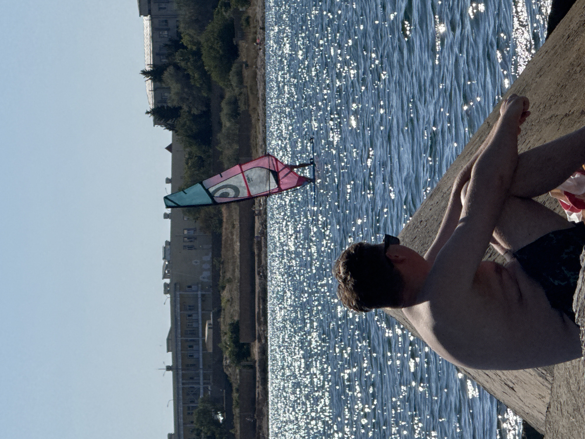 A medium shot captures a man sitting on a concrete structure by the sea on a sunny day in Sevastopol, Ukraine. The man, facing away from the camera and towards the water, wears dark swim trunks and sunglasses. His arms are resting on his knees, and his torso is bare.
The sea, a deep blue, is covered in sparkling highlights from the sun's reflection. In the distance, a windsurfer with a pink and teal sail navigates the water. Across the bay, a long, light-colored building complex stands against a clear blue sky, bordered by trees. The structure the man sits on appears to be part of a breakwater or pier, made of rough, weathered concrete.
