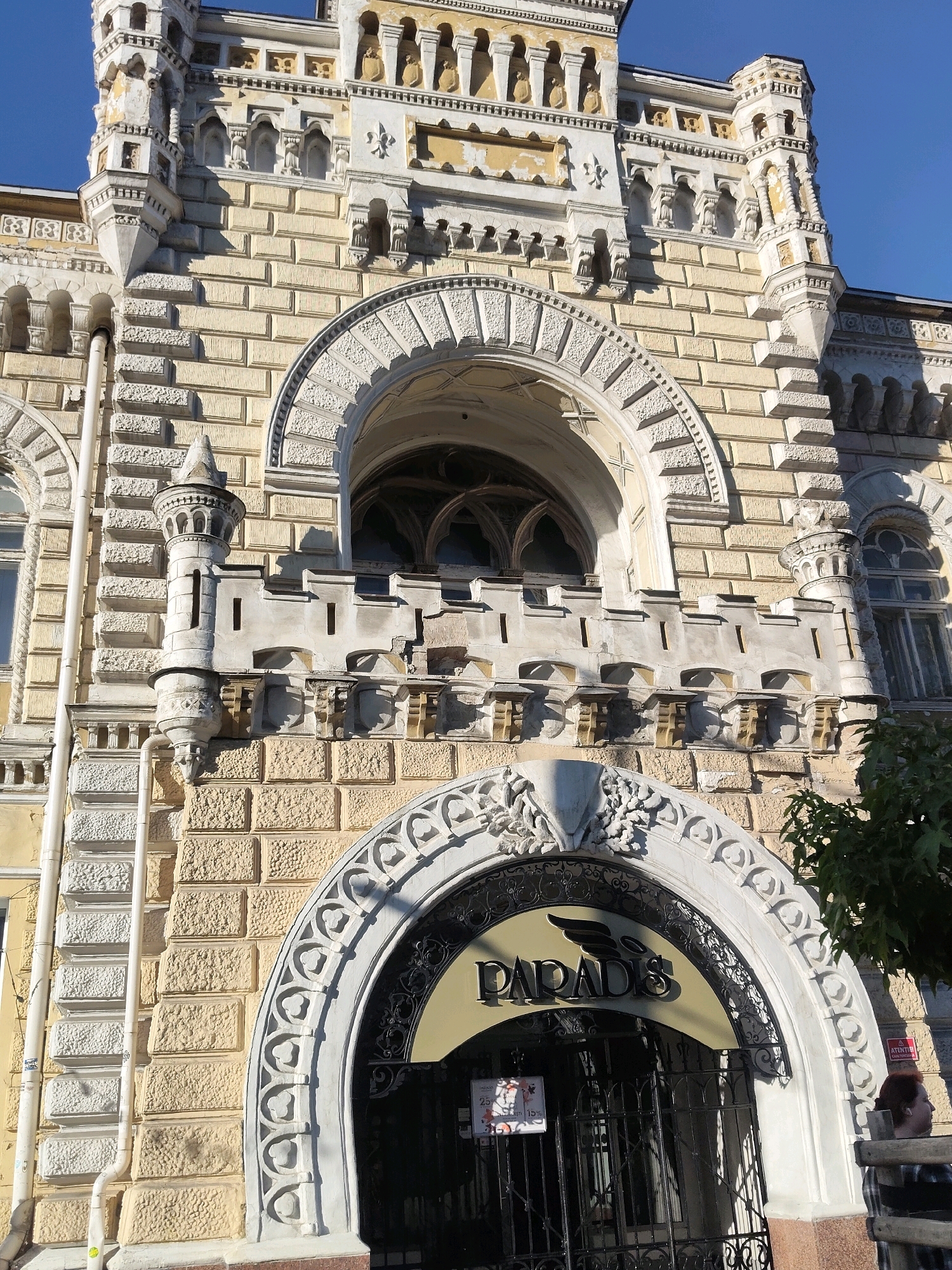 A low-angle, full-shot captures the ornate facade of a building on a clear, sunny day. The building is constructed from light yellow brick with white, intricate architectural details, including arched windows, decorative carvings, and turrets. A large, arched entrance with black metal gates is prominent in the lower center of the frame, featuring a sign that reads "PARADIS" with a winged emblem. To the right of the entrance, a person with reddish-brown hair is partially visible, looking away from the camera. A lush green tree branches into the upper right corner of the image. The sky above is a clear, vibrant blue.