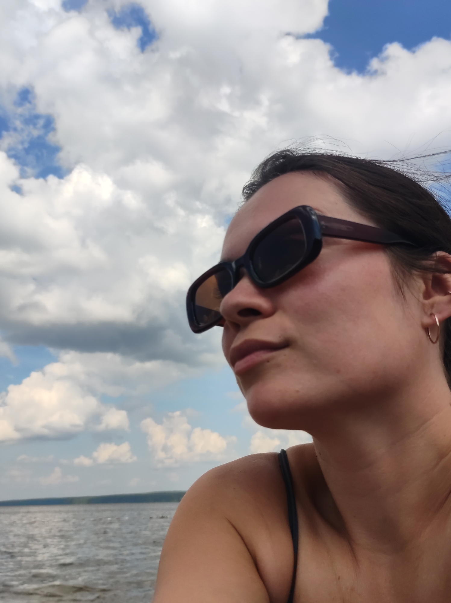 An outdoor, low-angle, medium shot captures a woman from the shoulders up, facing towards the right of the frame. She wears dark-rimmed sunglasses that obscure her eyes, and a subtle smile plays on her lips. Her dark hair is windswept. A small, gold hoop earring adorns her right ear. The background consists of a bright blue sky filled with fluffy white clouds. Below the sky, a body of water stretches to the horizon, bordered by a distant treeline. The image appears to be taken near a body of water in Chaikovsky, Russia.