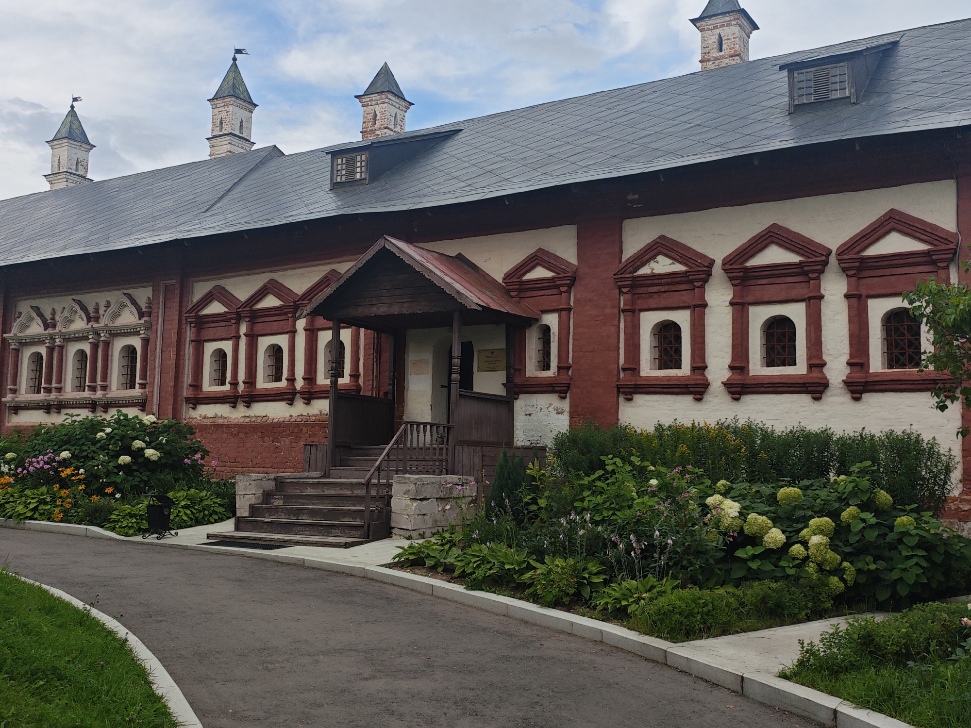 The image shows the Savvino-Storozhevsky Monastery in Zvenigorod, Russia. The main subject is a long, two-story building with a red brick lower level and a white-plastered upper level. The roof is dark gray and sloped, with several dormer windows. The building features numerous decorative elements, including arched window frames with red trim and ornate carvings above them on the upper level. Several pointed towers with flags rise from the roofline.
In front of the building is a landscaped garden with a variety of plants, including large bushes with white flowers, smaller pink and orange flowers, and dense green foliage. A paved walkway runs in front of the garden, separating it from a asphalt path that curves to the left. Wooden steps lead up to an entrance in the center of the building, which is covered by a wooden porch with a red roof. A metal trash can is visible in the garden. The sky is partly cloudy. There are no people visible in the image.