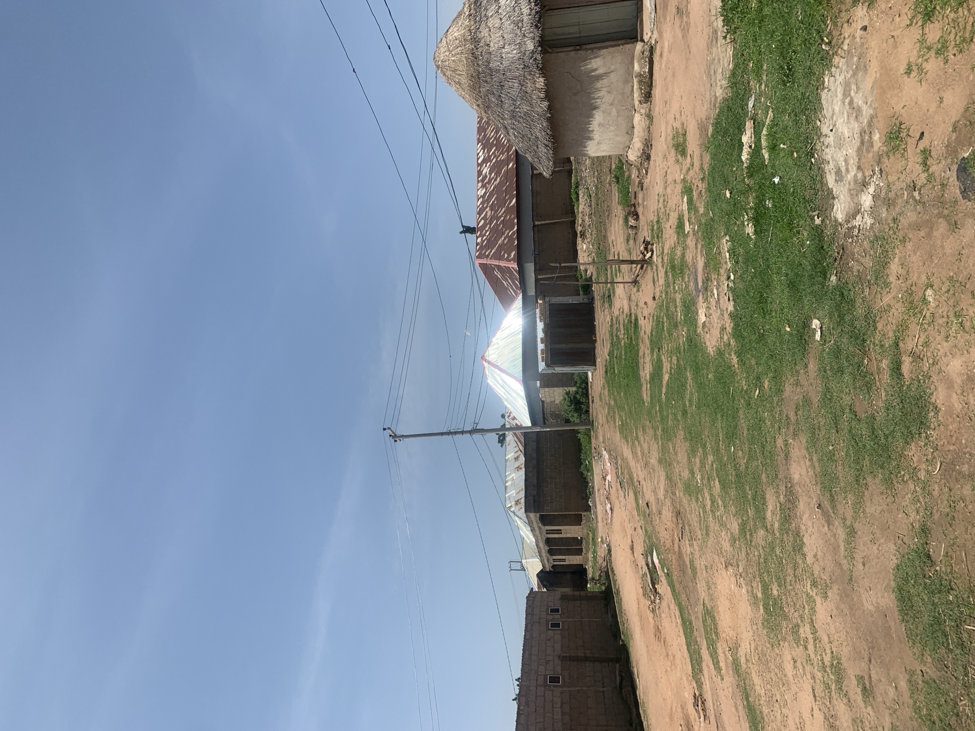 The image depicts a dusty, rural settlement in Jauro Yinu, Nigeria, under a clear blue sky. The foreground shows a dirt path with patches of sparse green grass and scattered debris. To the left, a low brick building with small square windows stands. In the mid-ground, several structures with corrugated metal roofs are visible, some appearing unfinished or partially constructed. A prominent, large tent with a white and green patterned canopy is set up in the center. To the right, a traditional round hut with a thatched roof is present. Utility poles and electrical wires crisscross the sky above the settlement, indicating the presence of electricity. The overall scene suggests a community in development or experiencing a temporary event given the tent. No people are explicitly visible in the frame.