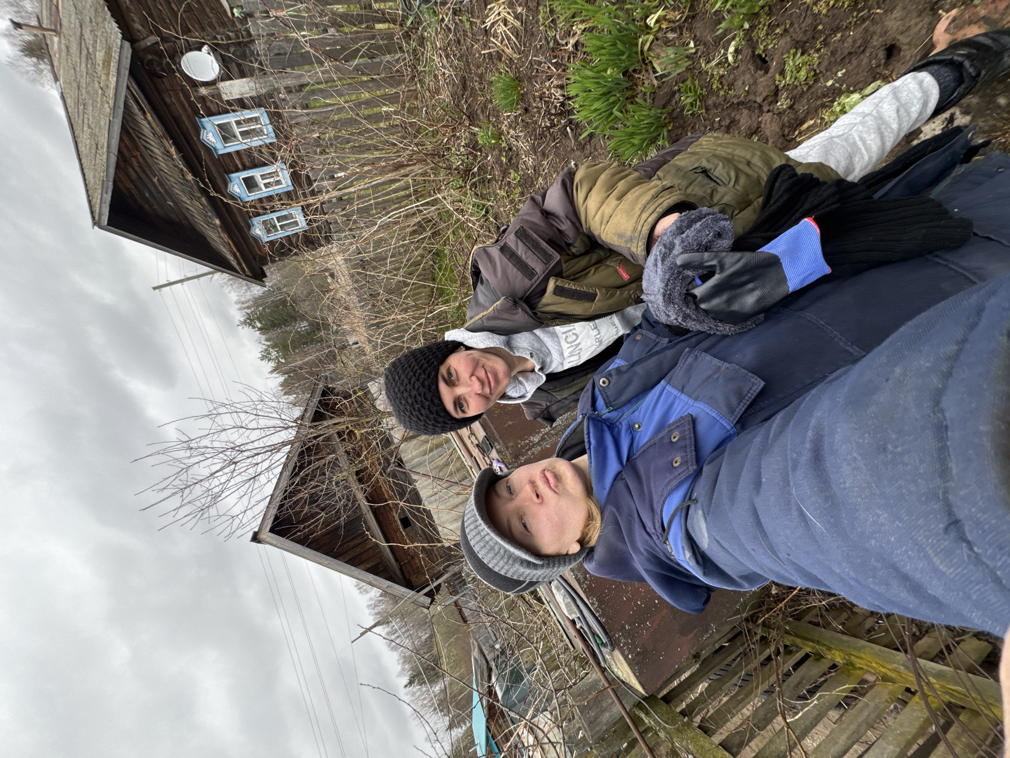 Two individuals are captured in a selfie-style photograph outdoors, likely in a rural or village setting in Вассята, Russia. The image, originally rotated sideways, shows them looking directly at the camera.
On the left, one person wears a black knit beanie, a grey hooded sweatshirt (with partial text "LANCH" visible), and a dark green or brown heavy jacket. They have a calm expression with a slight smile. Beside them, another person wears a grey and black striped beanie, a blue and black work-style jacket, and blue trousers. This individual also looks towards the camera with a neutral gaze. Black gloves and a grey fur-lined scarf are held by one of the individuals.
In the background, a rustic wooden house with a pitched roof is visible. The house features three windows with distinctive decorative blue frames (nalichniki) and a white round dish attached to its exterior. Surrounding the subjects are bare tree branches, dry undergrowth, patches of green grass, and wooden fences, indicative of late autumn, winter, or early spring. The sky is overcast, suggesting a cloudy day. The heavy clothing worn by both individuals further points to cool or cold weather conditions.