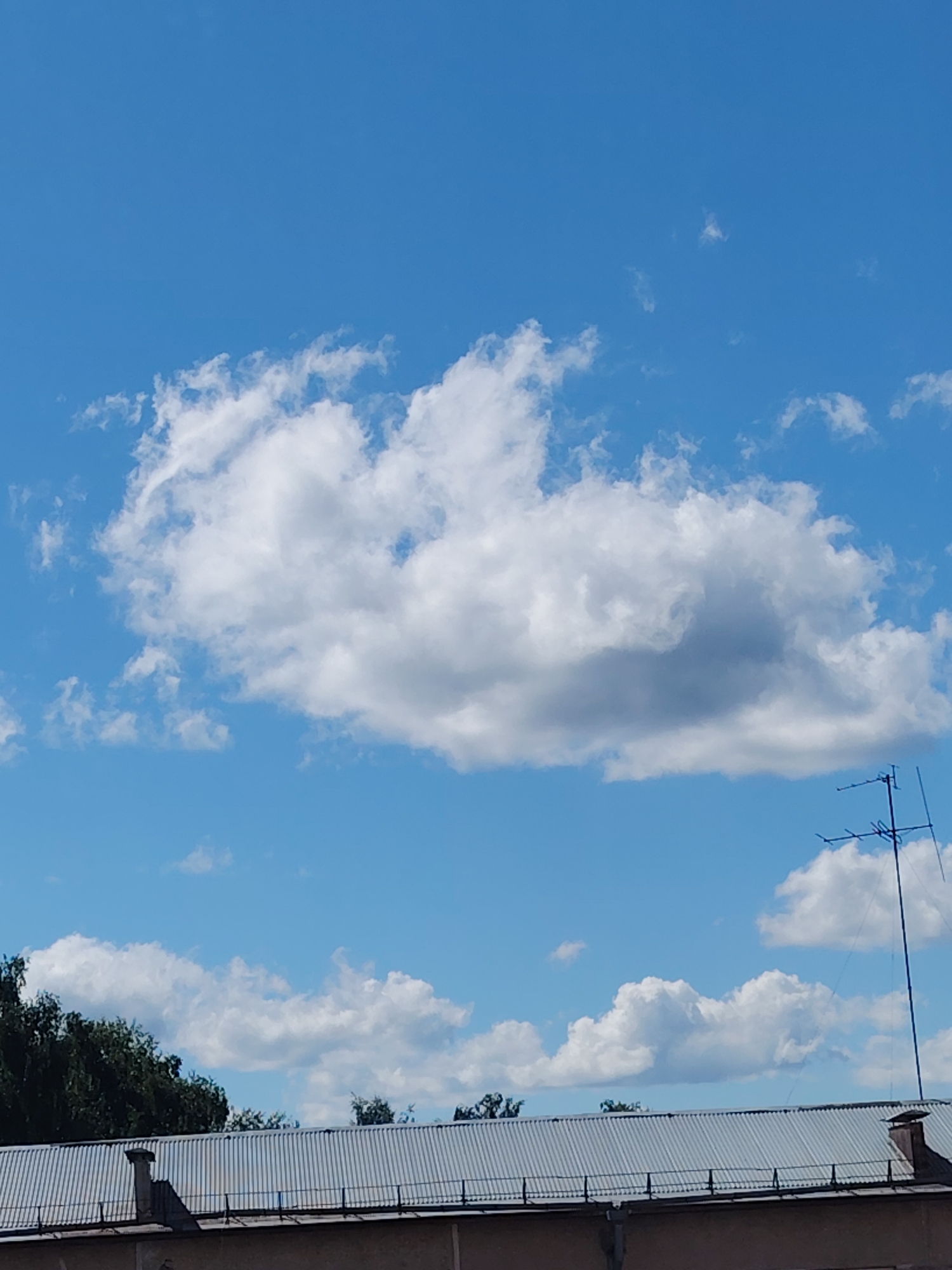 The image captures a clear blue sky with several white, fluffy clouds. In the foreground, a corrugated metal roof of a building is visible, extending across the bottom of the frame. Trees with green foliage can be seen behind the roof on the left side of the image. To the right, a television antenna stands tall against the sky. The scene appears to be an outdoor view from a city, possibly Konakovo, Russia, on a sunny day.