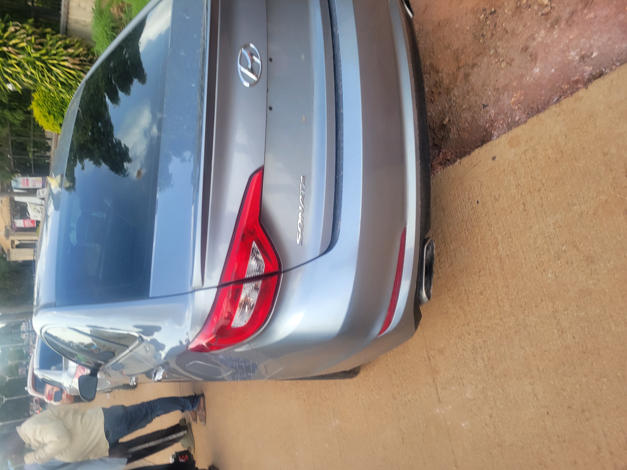 This is a vertical, eye-level, outdoor shot of the rear of a silver Hyundai Sonata sedan. The car is parked on a reddish-brown dirt and concrete surface, with some green foliage and a white building visible in the background. The sunlight suggests it is daytime. The car's taillights are red and the trunk has a chrome Hyundai logo and the "SONATA" badge. The car appears to be in good condition, with no visible damage. A person in a light-colored jacket and dark pants is standing in front of the car, partially obscured. Another person in dark clothing is also visible, possibly walking by. The overall scene is casual and everyday, suggesting the car is parked on a street or in a driveway. There is no discernible text in the image.