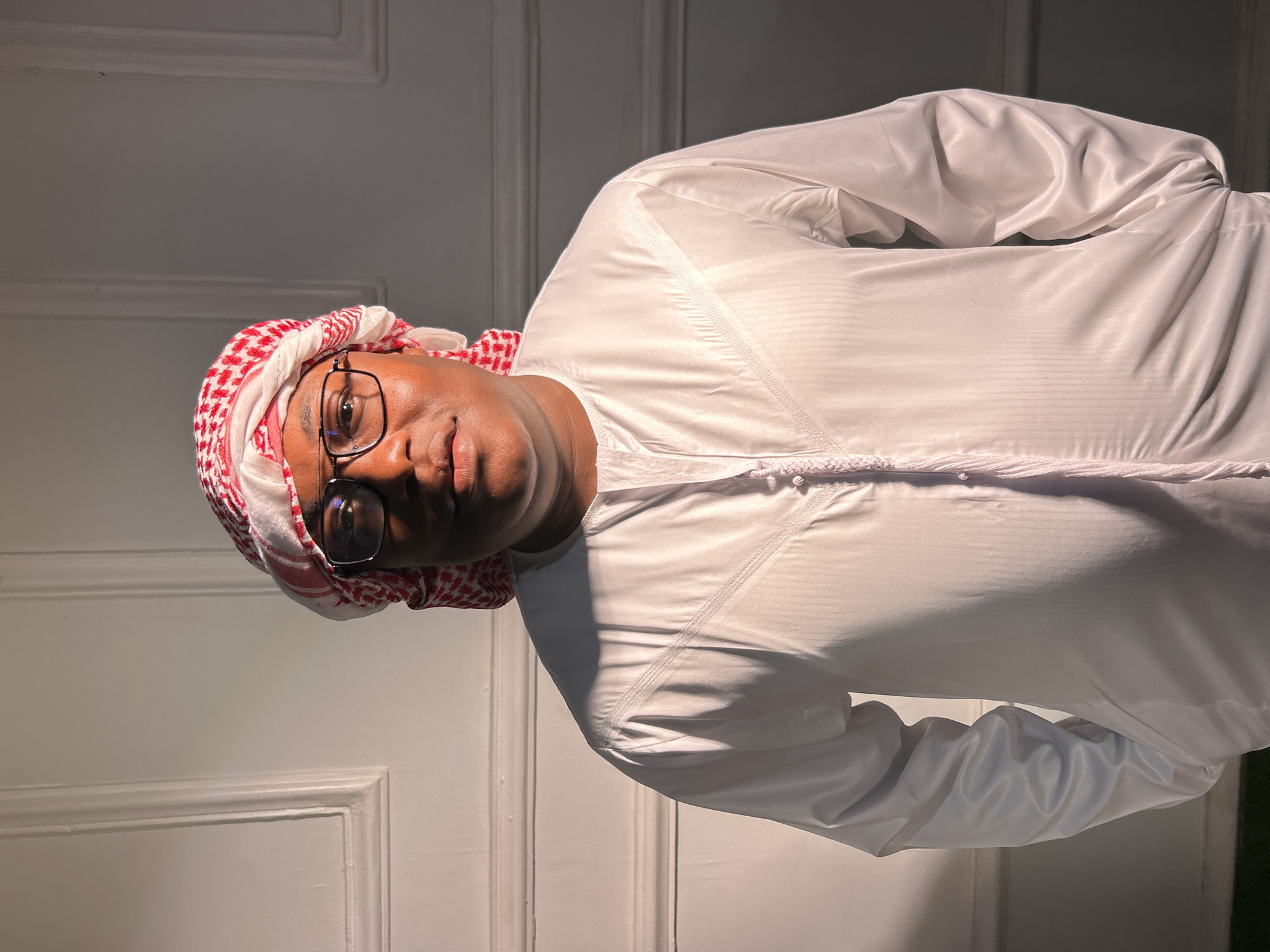 A man is depicted wearing traditional Middle Eastern attire, including a white thobe and a red and white checkered keffiyeh. He is also wearing glasses with a rectangular frame. The background appears to be a simple interior with a white paneled door. There is no evidence to connect this image to Jos, Nigeria.