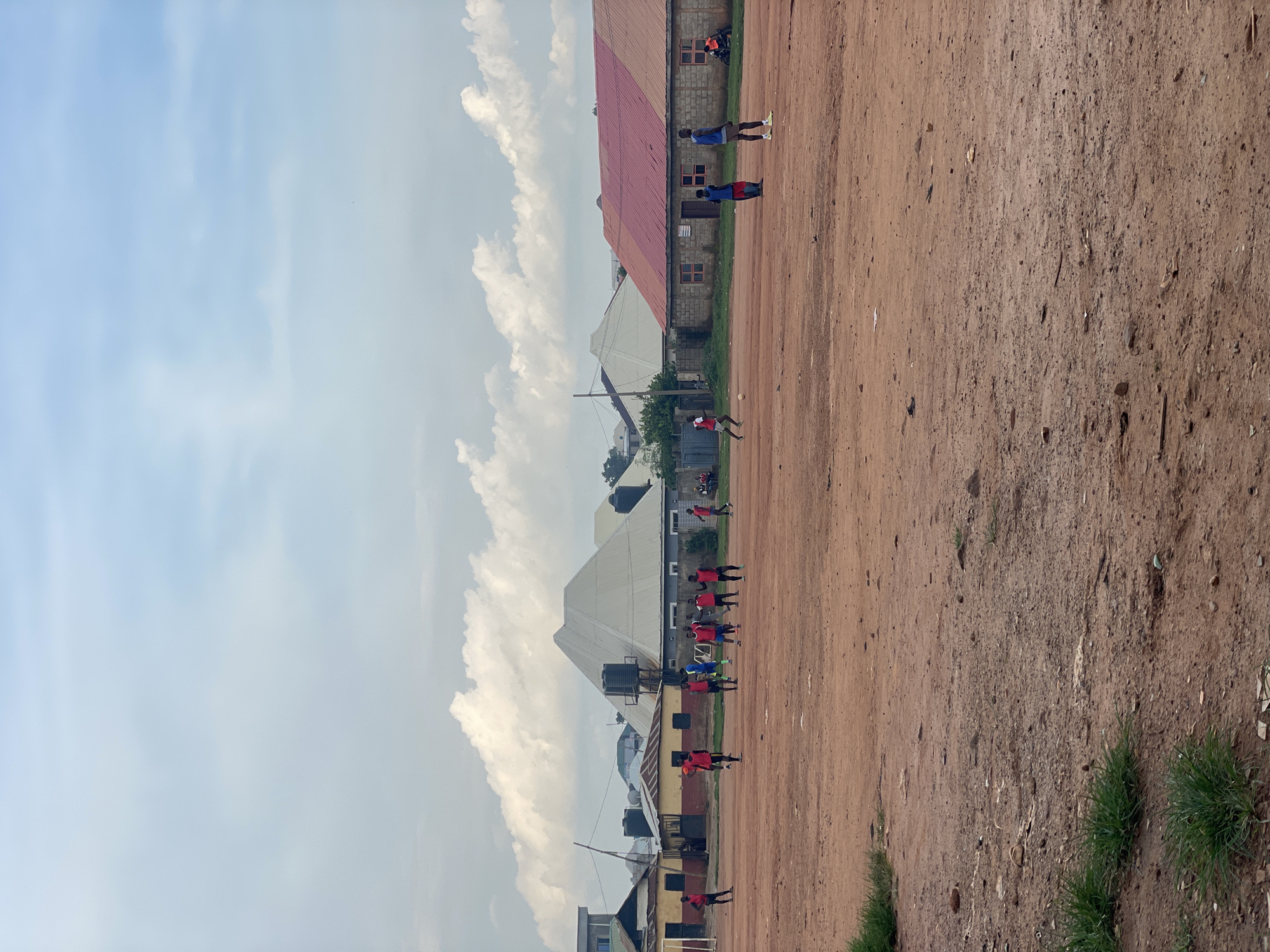 The image captures an outdoor scene in Kurunduma, Nigeria, where a group of young men are engaged in a game of football on a dirt field. The field is a reddish-brown color, with patches of dry grass and scattered debris. In the background, several buildings are visible, including structures with red and white roofs. A large, white, triangular-shaped building with a rounded apex stands out prominently. The sky is partly cloudy, with a mix of blue sky and white clouds. Several individuals are scattered across the field, some appearing to be actively playing football, while others are standing or observing. The men are dressed in casual clothing, predominantly in red and blue jerseys and shorts. The overall atmosphere suggests a typical community gathering for recreational activity in an urban or semi-urban setting in Nigeria.