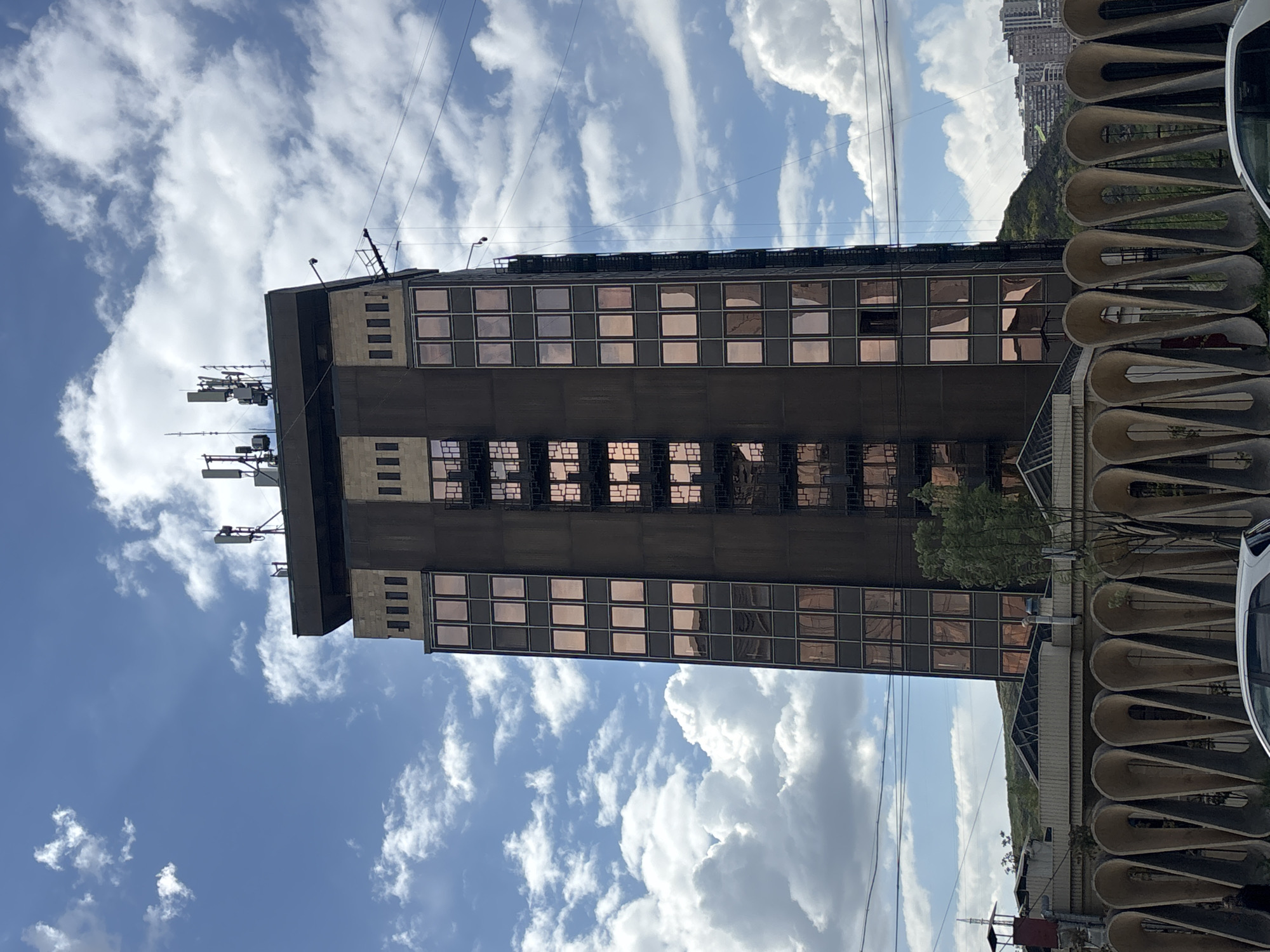 The image shows the Armenia Marriott Hotel Yerevan located in the city of Yerevan, Armenia. It is a tall, dark-colored building with multiple rows of windows reflecting the sky. It is located on Amiryan Street 4, Yerevan 0010, Armenia. A series of architectural, tear-drop shaped concrete structures are visible next to it on the right side, as well as a parked white vehicle. The sky is visible in the background with fluffy white clouds. There is a mountain range behind the building. There are antennas on top of the building.