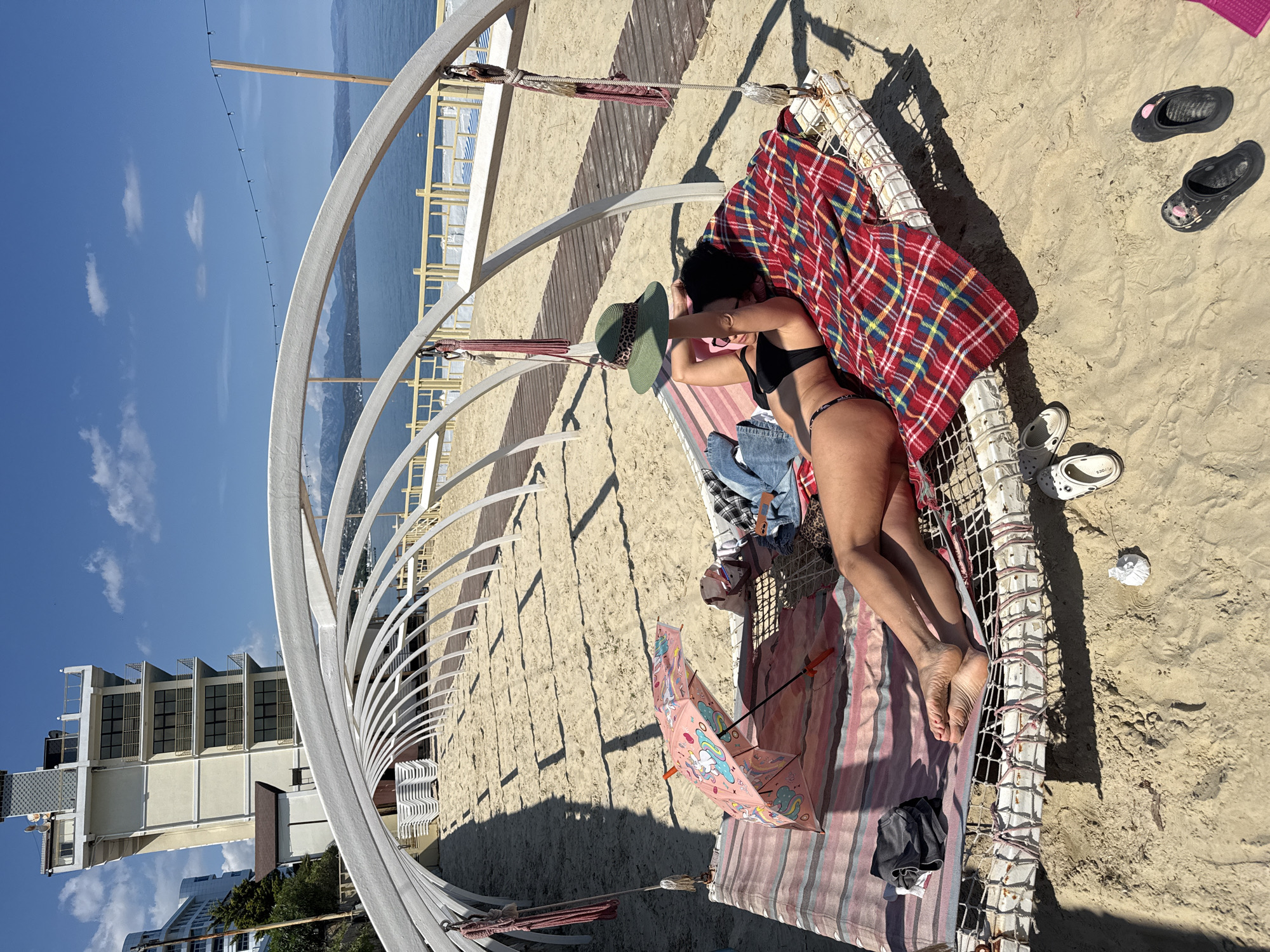 This media file captures a sun-drenched beach scene from an elevated perspective. The main subject is a person, likely female, lying face down on a large, net-like bed or hammock, covered partly by a red plaid blanket and a striped mat. She is wearing a black bikini and has a green wide-brimmed hat with a leopard print band near her head. A pair of jeans rests on the bed next to her.
The bed is situated on light-colored sand, partially enclosed by a prominent white arched architectural structure. Next to the bed on the sand is a vibrant pink umbrella adorned with cartoon unicorns and other playful designs. Nearby, a pair of black sandals and a pair of white Crocs-style shoes are visible on the sand, with the brand name "CROCS" faintly legible on one sole.
In the background, a calm blue body of water extends to the horizon, with distant mountains along the far shore. The sky is clear and bright blue with a few scattered white clouds. To the left, several multi-story buildings, typical of a coastal resort or town, can be seen. A wooden boardwalk traverses the sand. The strong sunlight casts distinct shadows across the sand and objects, indicating it is daytime. The overall impression is one of relaxation and leisure by the sea.