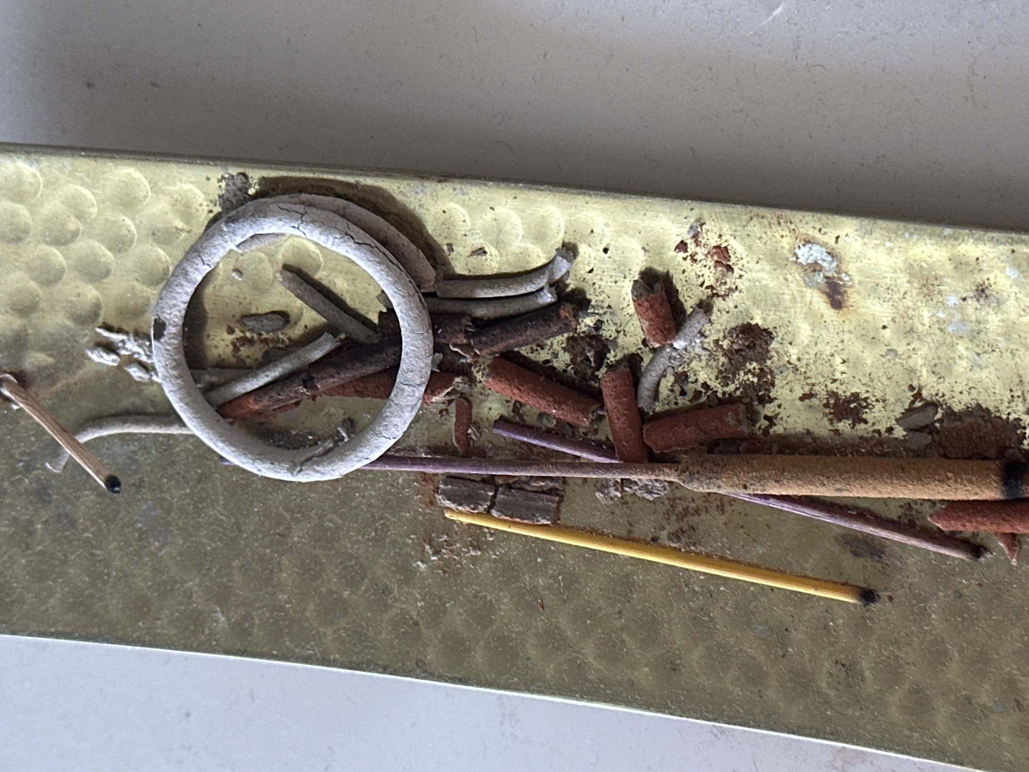 The image shows a close-up, overhead view of a brass incense holder with various burnt and unburnt incense sticks, ash, and a ring of incense on a textured, gold-colored surface. The incense holder is rectangular and appears to have a patterned surface. There are several broken pieces of incense sticks, some red and some brown, scattered around. A single yellow incense stick is positioned diagonally, and two purple incense sticks are leaning against it. A white, coiled incense ring is also present, with ash and debris around it. The background is a plain, light-colored surface. The image is devoid of any context that would indicate its location in Nevinnomyssk, Russia, or any other specific city. There are no people, actions, or interactions depicted.