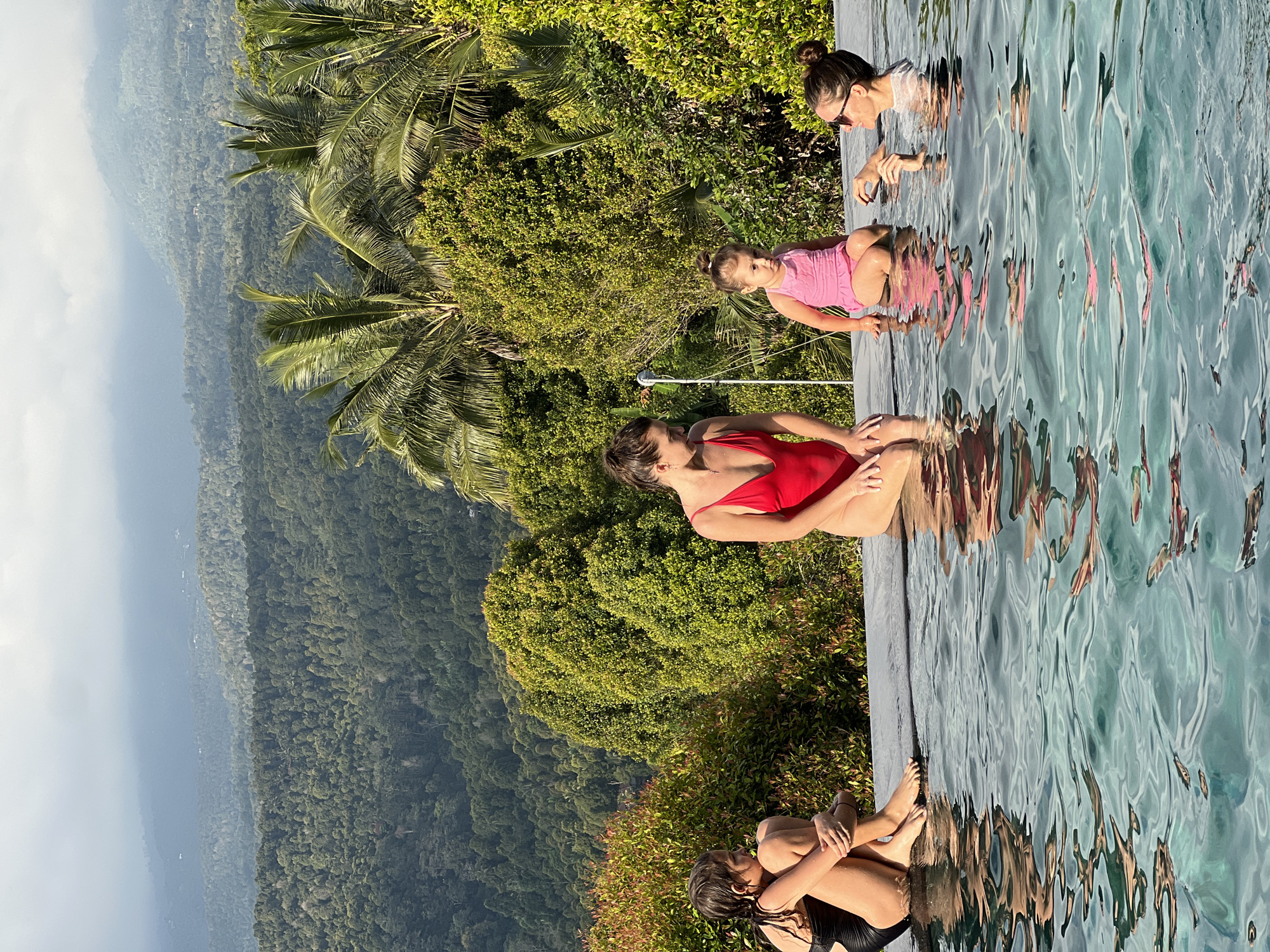 In Busungbiu, Indonesia, four individuals are present at the edge of an infinity pool. On the far left, a young person in a black swimsuit sits with their legs crossed, looking to the left. Next to them, a woman in a red swimsuit is seated, facing right. A young child in a pink swimsuit is positioned in the center, looking slightly to the left. On the right, a woman wearing sunglasses and a white top partially submerged in the water is looking at the child. The background features lush green trees and mountainous terrain under a cloudy sky.