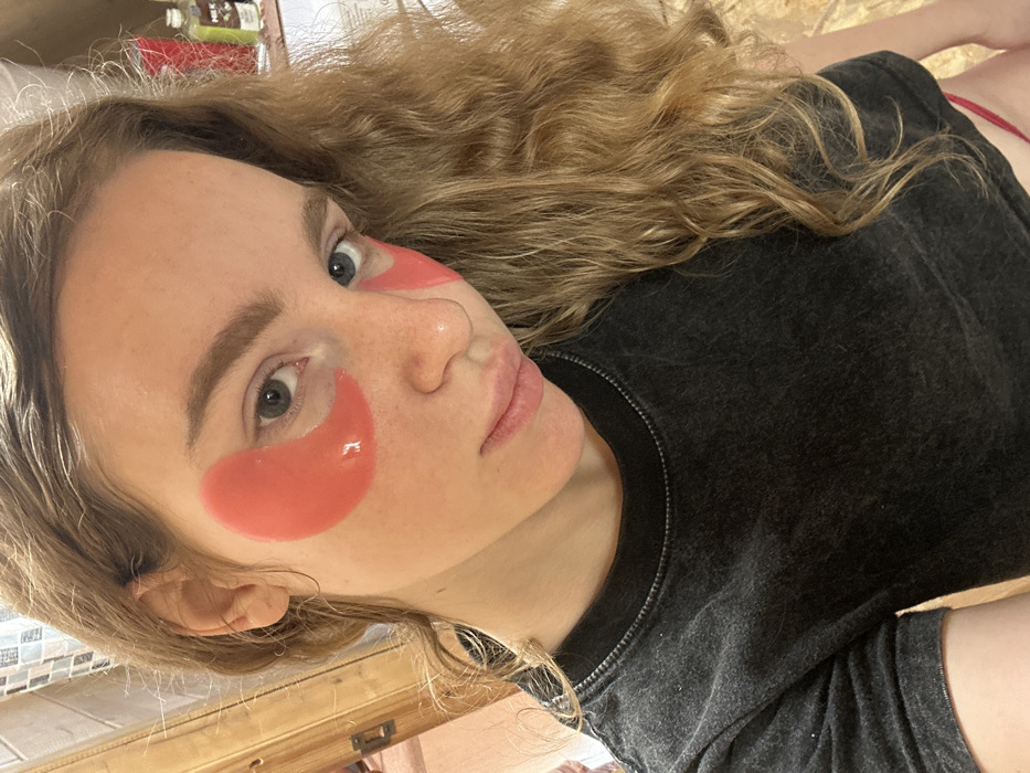 A single female subject is depicted in a close-up, chest-up indoor selfie, looking directly towards the camera. She has long, wavy, light brown hair with blonde highlights, fair skin with visible freckles across her nose and cheeks, light-colored eyes (appears blue/grey), and defined eyebrows. Two translucent, coral-pink hydrogel under-eye patches are applied beneath each eye, covering the orbital area. Her lips are slightly parted. She is wearing a dark grey or charcoal-colored short-sleeved t-shirt with a ribbed crew neck and subtle white stitching along the collar. A thin red strap, possibly from an undergarment, is visible at the bottom right edge of the frame.
The background elements suggest an interior residential setting. To the left, a portion of a light brown wooden door or cabinet is visible, adjacent to a section of a wall tiled with light-colored, possibly white or beige, small rectangular tiles. To the right, various bottles are arranged on a countertop or shelf, including a red bottle, a dark green bottle labeled "MILK" (partially visible), and other indistinct containers, indicating a potential bathroom or dressing area. No specific identifying features confirming the location as Vasilyevo, Russia, are discernible within the image.