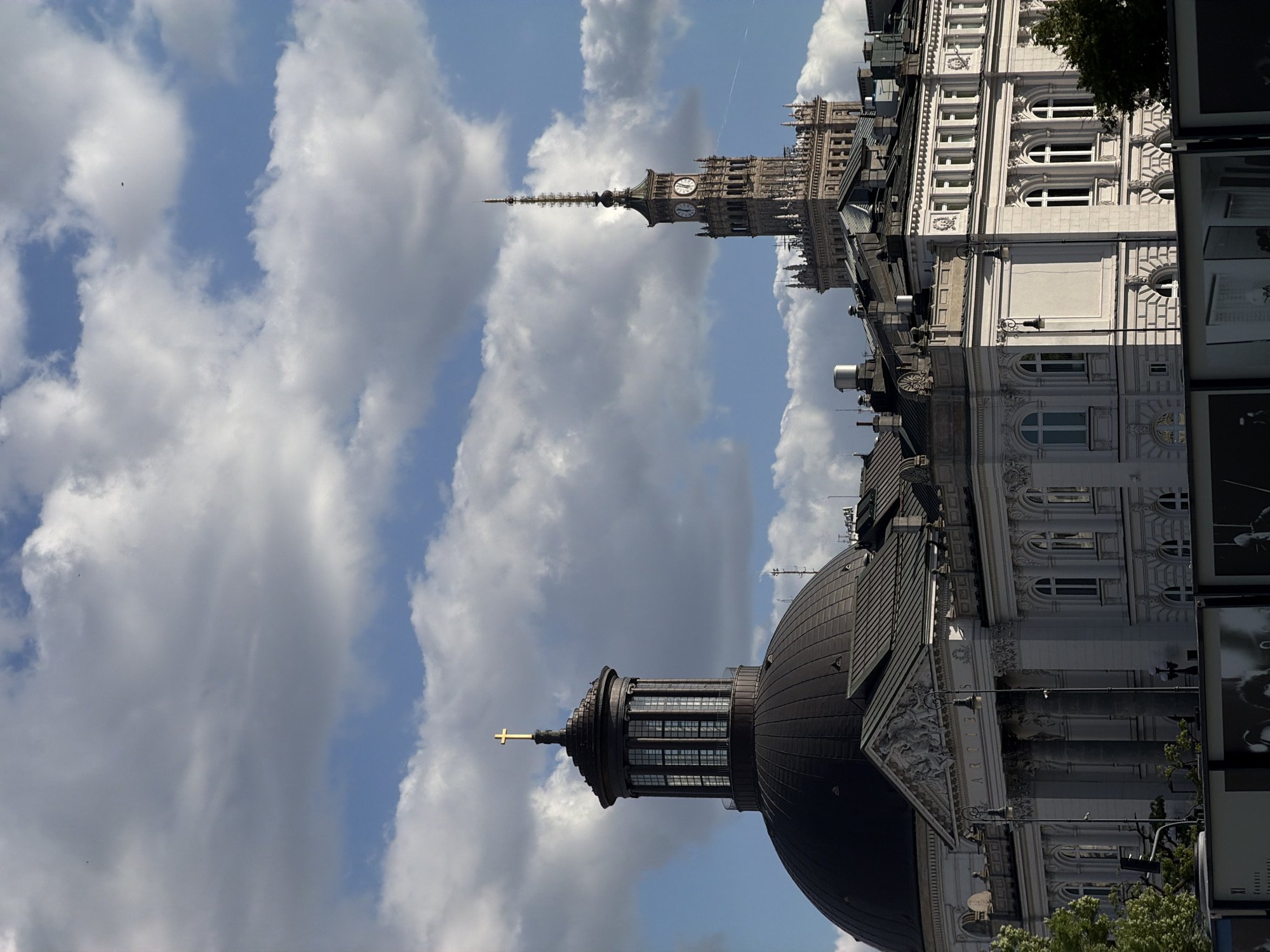 The image shows a view of buildings and the sky in an urban setting. In the foreground, there is a large dome structure with a cross on top, likely part of a significant building. Behind the dome, more buildings with architectural details and windows can be seen. In the background, a tall, ornate tower with a clock face is visible. The sky is filled with clouds, and it appears to be a partly sunny day. The location appears to be Unknown, Unknown.