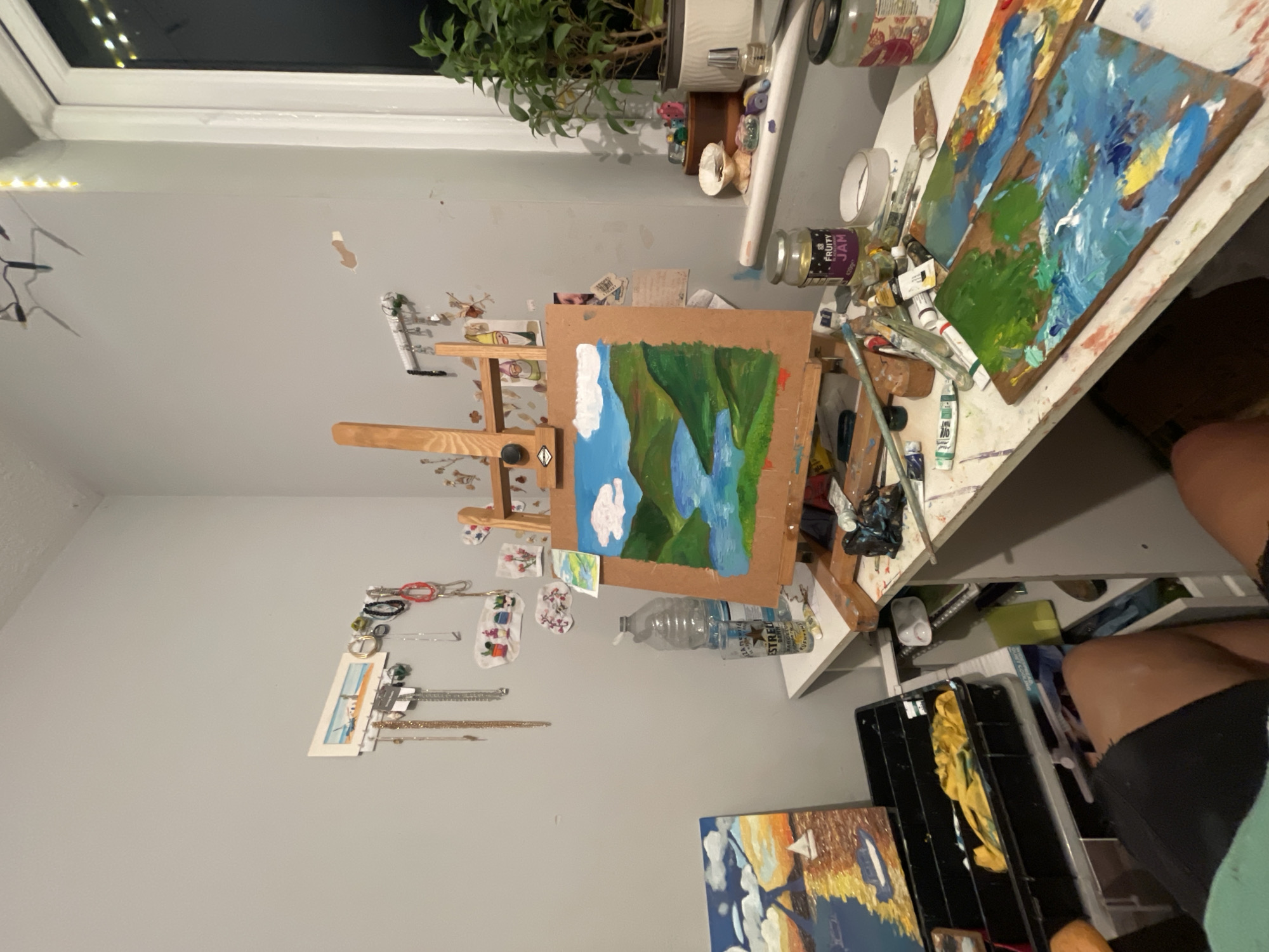 The image shows an artist's studio. An easel holds a painting in progress depicting a landscape with mountains, trees, and a body of water, likely done in oil or acrylic paints. The painting is displayed on a wooden easel. The studio space is cluttered with painting supplies, including palettes covered in paint, brushes, tubes of paint, jars, and other art-related items. There's also a water bottle on a shelf next to the easel. Other artwork is visible in the background. There is no indication in the image of the city of Maidstone, United Kingdom. There are no people visible in the image, apart from a glimpse of a person's leg and clothing at the right side of the frame.