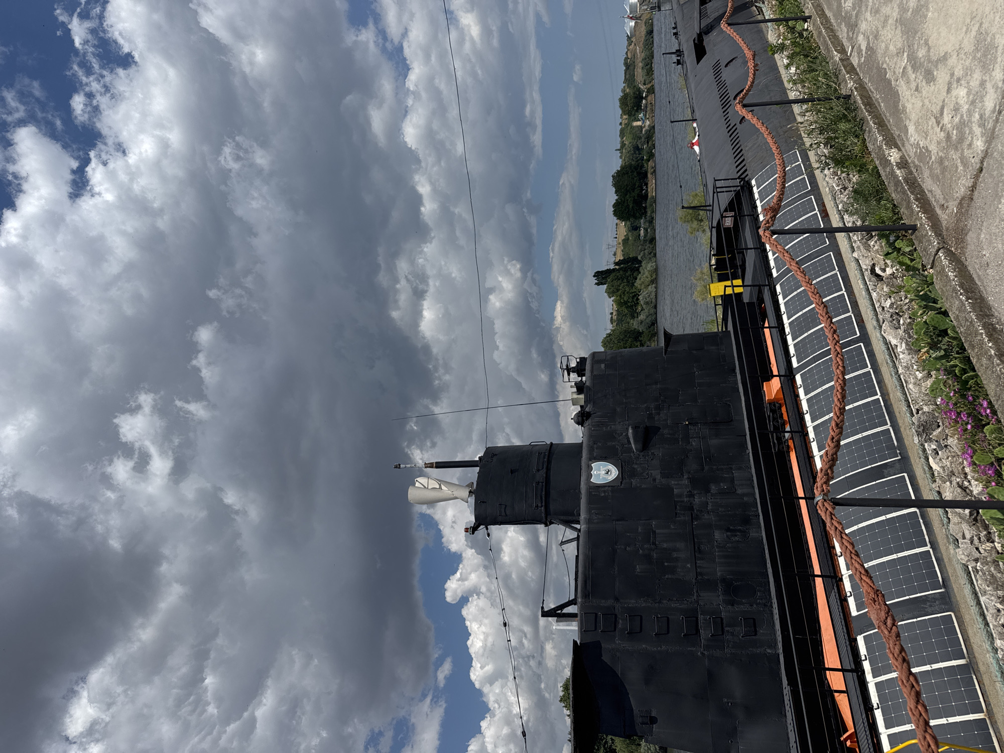 An outdoor shot captures a black submarine, likely a museum exhibit, docked on the shore of a river under a dramatic, cloudy sky. The submarine's conning tower, featuring a white exhaust stack and antenna, is prominent. The side of the submarine displays a crest or insignia and a series of square panels, possibly solar panels, running along its hull. A thick, reddish-brown rope cordons off the area in front of the submarine, which is bordered by rocky terrain and some low-lying purple flowers. In the background, across the river, a tree-lined shore is visible, with a few buildings and utility poles on the distant horizon. The scene is located in Beloslav, Bulgaria.