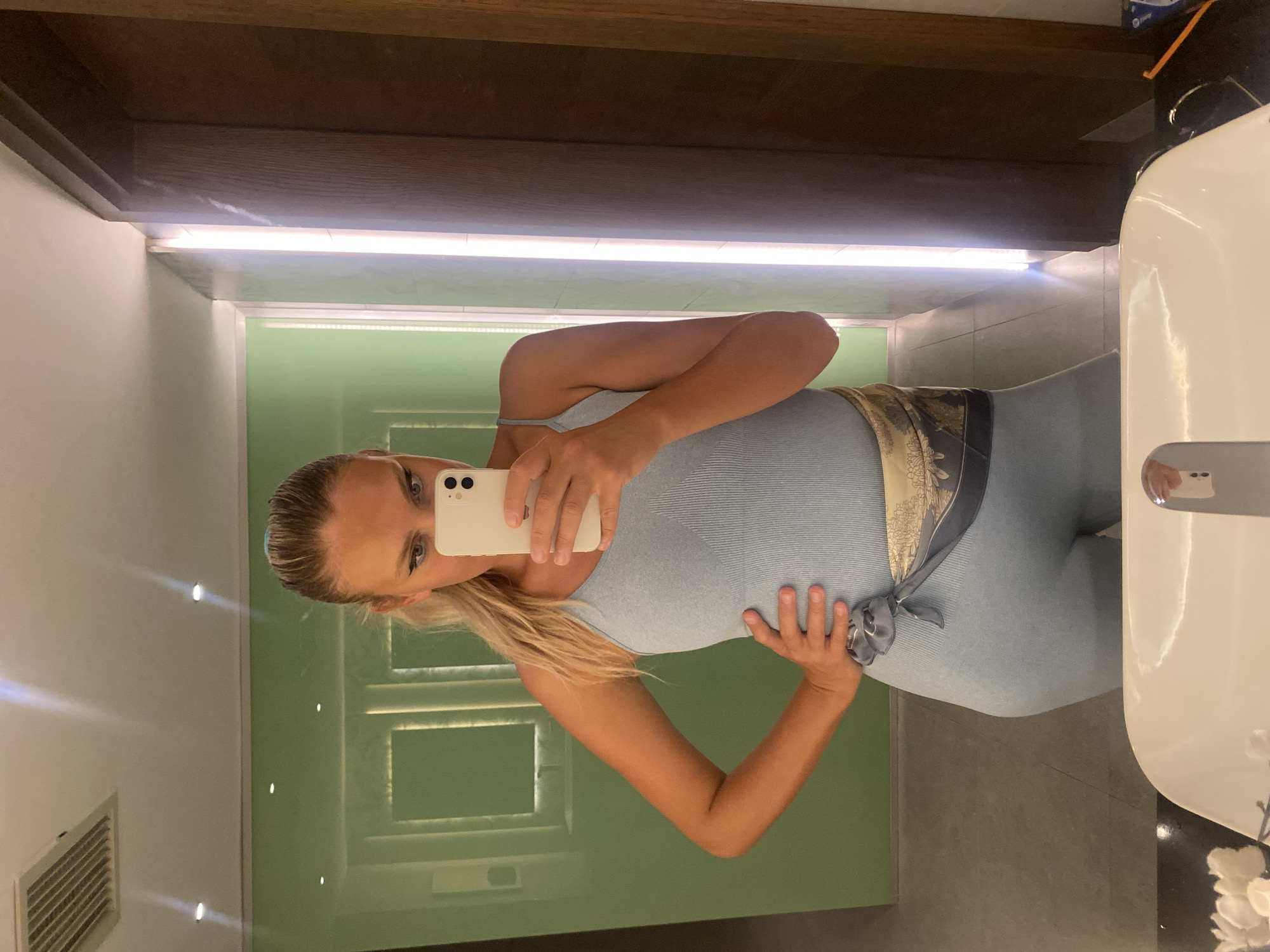 A woman with blonde hair pulled back in a ponytail takes a mirror selfie in what appears to be a bathroom. She is wearing a light blue ribbed jumpsuit and has a scarf tied around her waist. Her left hand is on her hip, and she is holding a white smartphone in her right hand. The background shows a green tiled wall with recessed lighting and a wooden doorframe. The scene is lit by bright artificial lights, casting reflections on the mirror and surrounding surfaces. There are no specific landmarks or indications of the city of Aksu, Turkey, visible in the image.