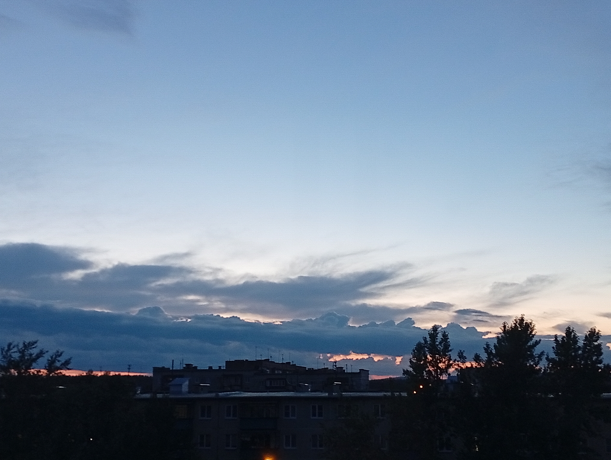 This image depicts a twilight scene over a residential area, likely Kyshtym, Russia. The sky is a gradient of pale blue at the top, transitioning to a lighter hue near the horizon, indicating either sunrise or sunset. Wispy, dark clouds are scattered across the lower portion of the sky, with some illuminated by a faint orange glow from the setting or rising sun, suggesting a transitionary weather pattern.
In the foreground, the silhouettes of trees and buildings are visible. The buildings appear to be apartment blocks, common in Russian residential areas, with visible windows and antenna structures on the rooftops. The overall mood is calm and serene, characteristic of dusk or dawn. The lighting is dim, with the last vestiges of daylight illuminating the sky and casting the foreground elements into shadow. No people are discernible in the image. The image captures a quiet moment in an urban or semi-urban landscape at a time of day marked by subtle color shifts and diffused light.