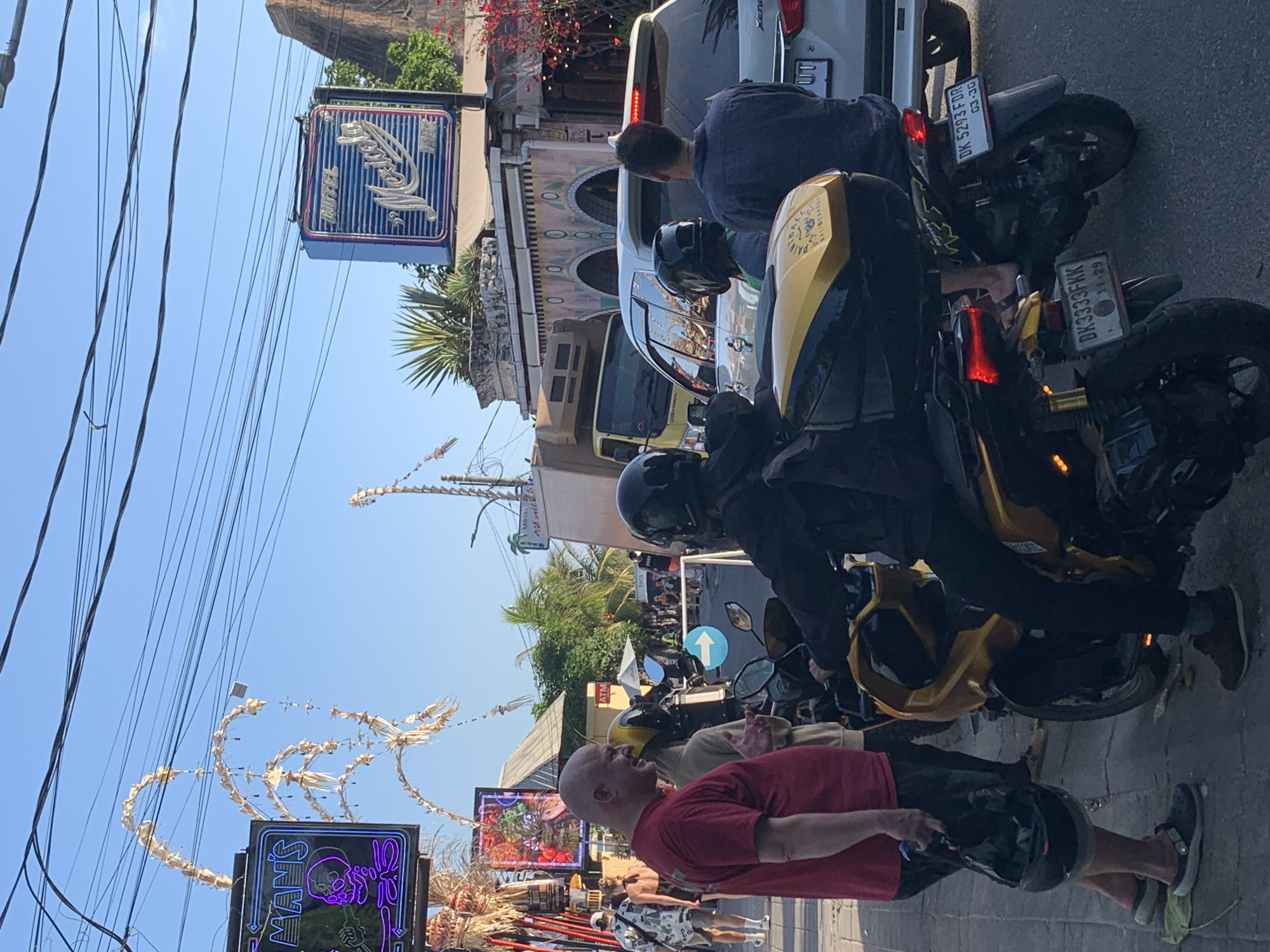 In North Kuta, Indonesia, a street scene unfolds with several individuals and vehicles. A bald man in a red t-shirt stands on the left, holding a black helmet. Next to him, a person in a light-colored hoodie sits on a yellow and black motorcycle, also wearing a helmet. Another person, wearing a black jacket and helmet, is seated on a similar motorcycle behind the first. A silver SUV is parked behind the motorcycles. In the background, a hotel sign reads "HOTEL Mexicola" and another sign displays "MAN'S" with a neon skull. The environment appears to be a street with overhead wires and tropical vegetation. The lighting suggests it is daytime, possibly late afternoon, with clear skies.