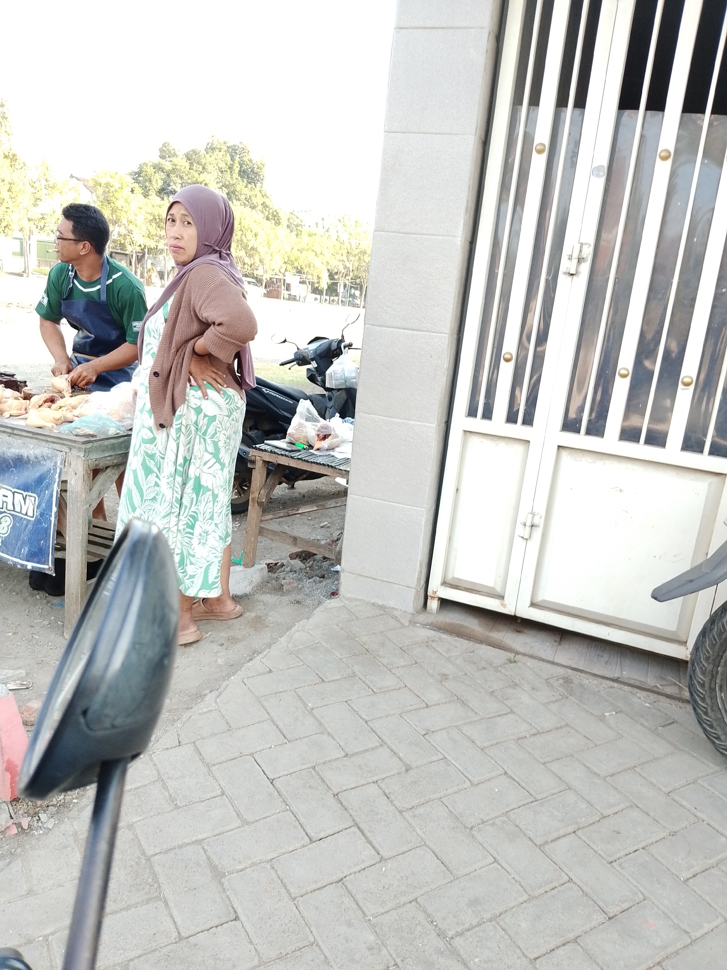 In Pasuruan, Indonesia, a scene unfolds at what appears to be an outdoor market or roadside stall. A man, wearing a green shirt and a dark blue apron, is focused on preparing poultry, with several raw chickens laid out on a wooden table. To his right, a woman stands, dressed in a hijab and a patterned dress with a cardigan. Her stance suggests she is either a vendor or an observer. Behind them, a motorcycle is parked, indicating a mode of transportation common in the area. The ground is a mix of dirt and paved brickwork. In the foreground, a motorcycle's rearview mirror is visible, suggesting the photo was taken from a parked motorcycle. The backdrop features trees and what looks like a building with a white, vertically slatted gate. The overall ambiance is that of a typical, bustling Indonesian street scene.