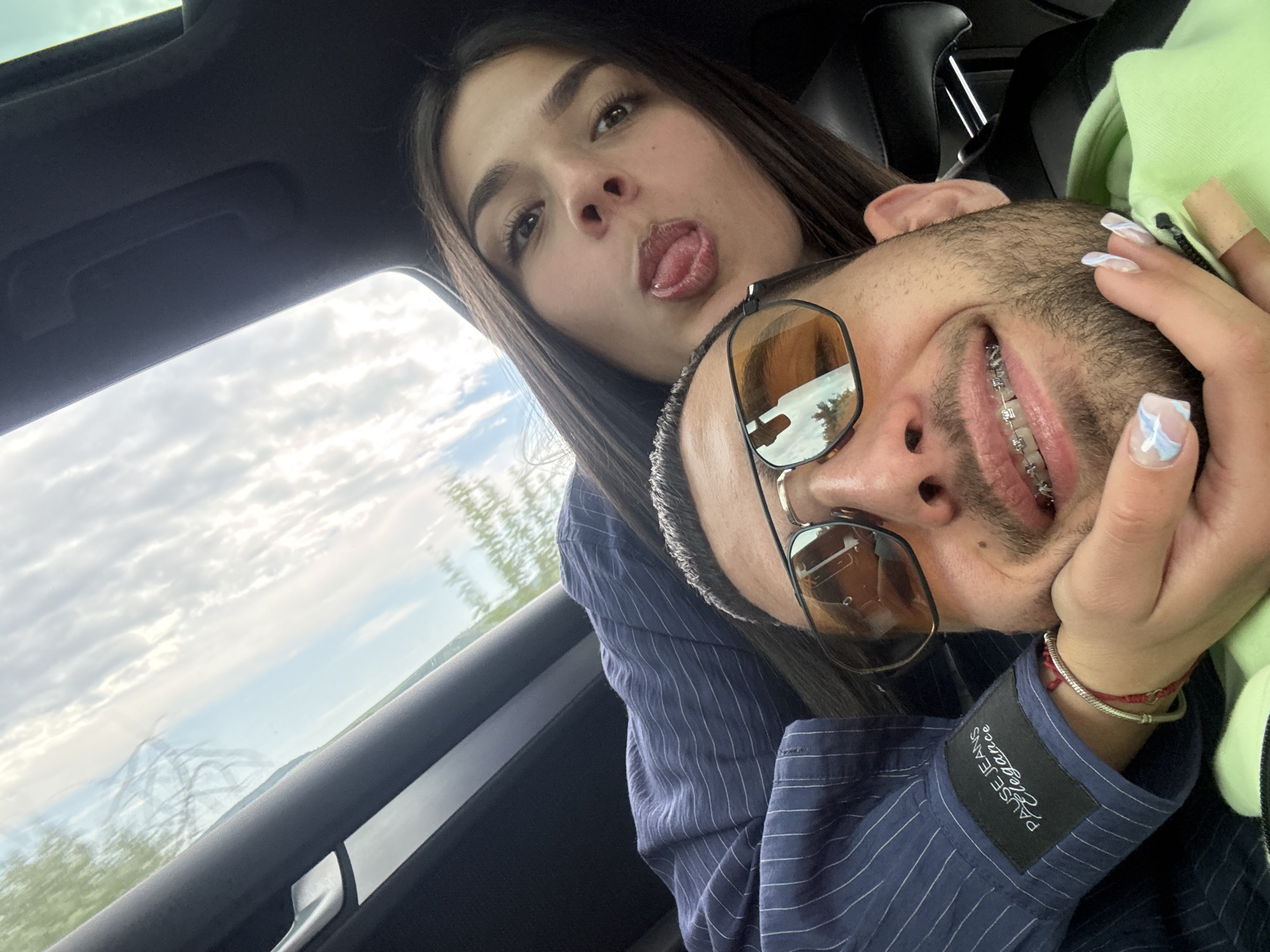 A young woman and man pose for a selfie inside a car. The woman sticks her tongue out. The man wears sunglasses and braces, and his head is supported by her hand. He wears a blue striped shirt with "PAUSE JEANS Elegance" label visible on the cuff. The car window shows a cloudy sky and foliage. The location is unlikely to be Polski Trambesh, Bulgaria, as the local environment does not align with the background visible.
