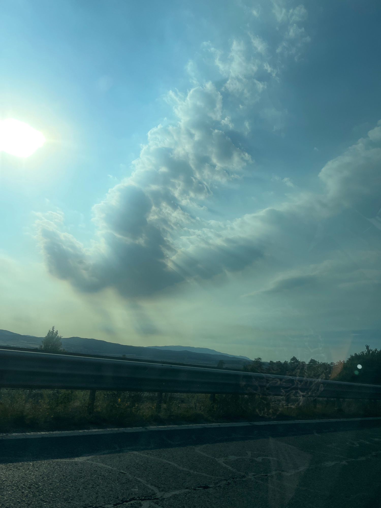The image captures a dramatic sky over the city of Ihtiman, Bulgaria. In the upper left quadrant, the sun shines brightly, its rays piercing through a vast expanse of blue sky. A prominent, wispy cloud formation dominates the center of the sky, appearing almost as if it is reaching towards the viewer. These clouds are illuminated by the sunlight, casting ethereal rays that create a sense of depth and movement.
Below the sky, a metal guardrail runs horizontally across the frame, separating the roadway from the landscape beyond. The roadway itself is paved and appears to be slightly cracked in places. Beyond the guardrail, a grassy verge is visible, dotted with some sparse vegetation. In the distance, a range of rolling hills forms the horizon line, shrouded in a hazy, greenish-blue light. The overall impression is one of a vast, open landscape under a dynamic and impressive sky. There are no people or specific actions depicted in the image. The environment suggests a roadside view, likely from a vehicle in motion, given the angle and perspective.