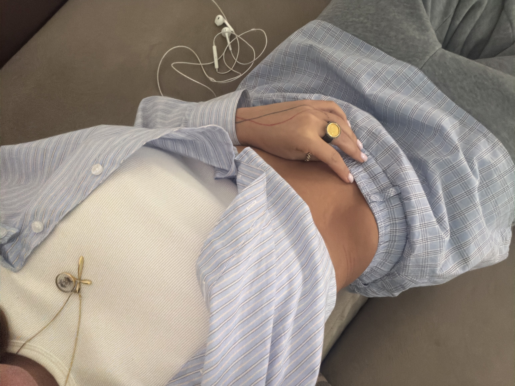 The image shows a person lying on a taupe-colored couch. The person is wearing a white tank top and a light blue striped button-down shirt. They have a gold pendant necklace on and are wearing rings on their fingers, including a large gold ring with a dark setting. Next to their hand, there are white wired earbuds. The couch is in a neutral-toned room. There is no indication of the city of Yerevan, Armenia.