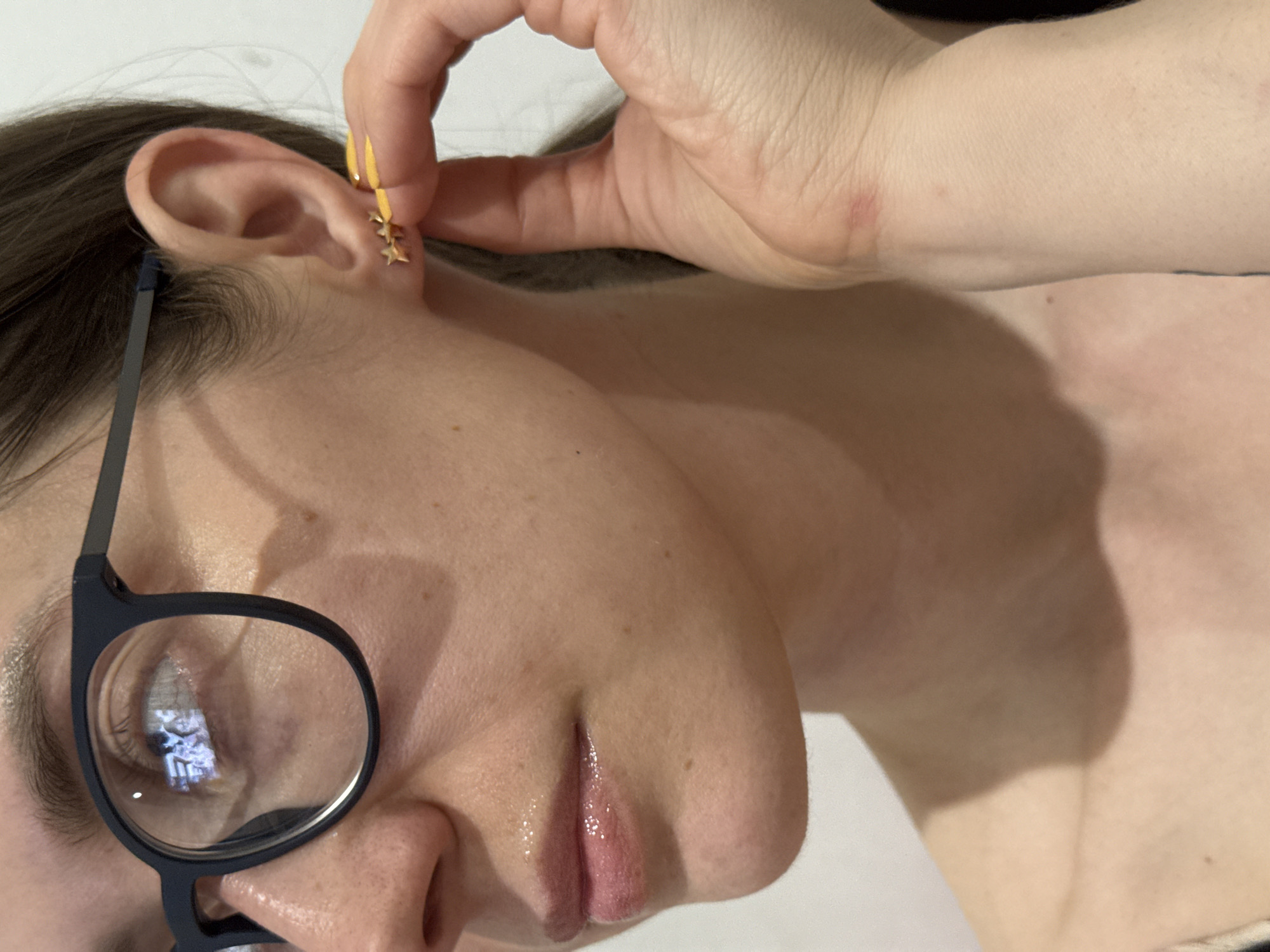 A person with fair skin, dark hair, and wearing eyeglasses is adjusting a gold-colored earring in their right ear. The earring has multiple star-shaped charms. The background is a plain white color. The scene appears to be indoors. There is no information to suggest this image was taken in Chaikovsky, Russia.