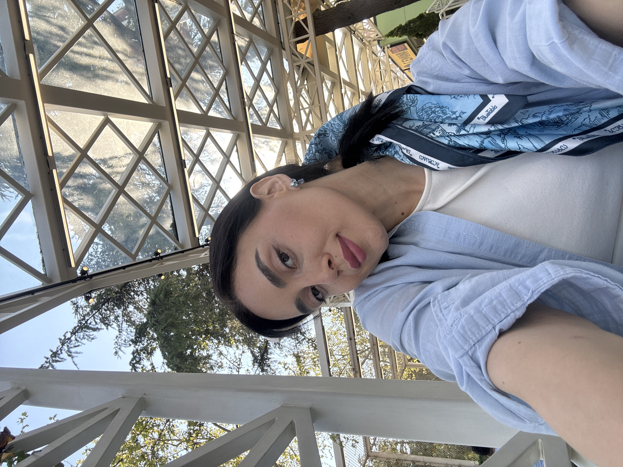 A selfie-style portrait captures a woman with dark hair and a playful expression, her head tilted, and tongue slightly extended. She wears small blue stud earrings, a white top, and a light blue collared shirt. A blue patterned scarf is draped around her, featuring illustrations of buildings and possibly landscapes. The scarf has visible text including "BlueTablo" and Georgian script that appears to read "ბლუ ტაბლო" (Blu T'ablo).
The setting is an airy, brightly lit space, likely a conservatory, veranda, or an enclosed outdoor dining area, characterized by a white structural framework and large diamond-paned windows. Through the windows, bright daylight is visible, along with green trees and a clear blue sky. Some sections of the window frames have vertical light strips integrated into them, and small round lights are attached to the overhead structure. A white railing is prominent in the foreground, indicating the woman is seated or standing near a barrier. The overall impression is a sunny day in an elegant, semi-outdoor environment, potentially in Tbilisi, Georgia, as suggested by the Georgian script.