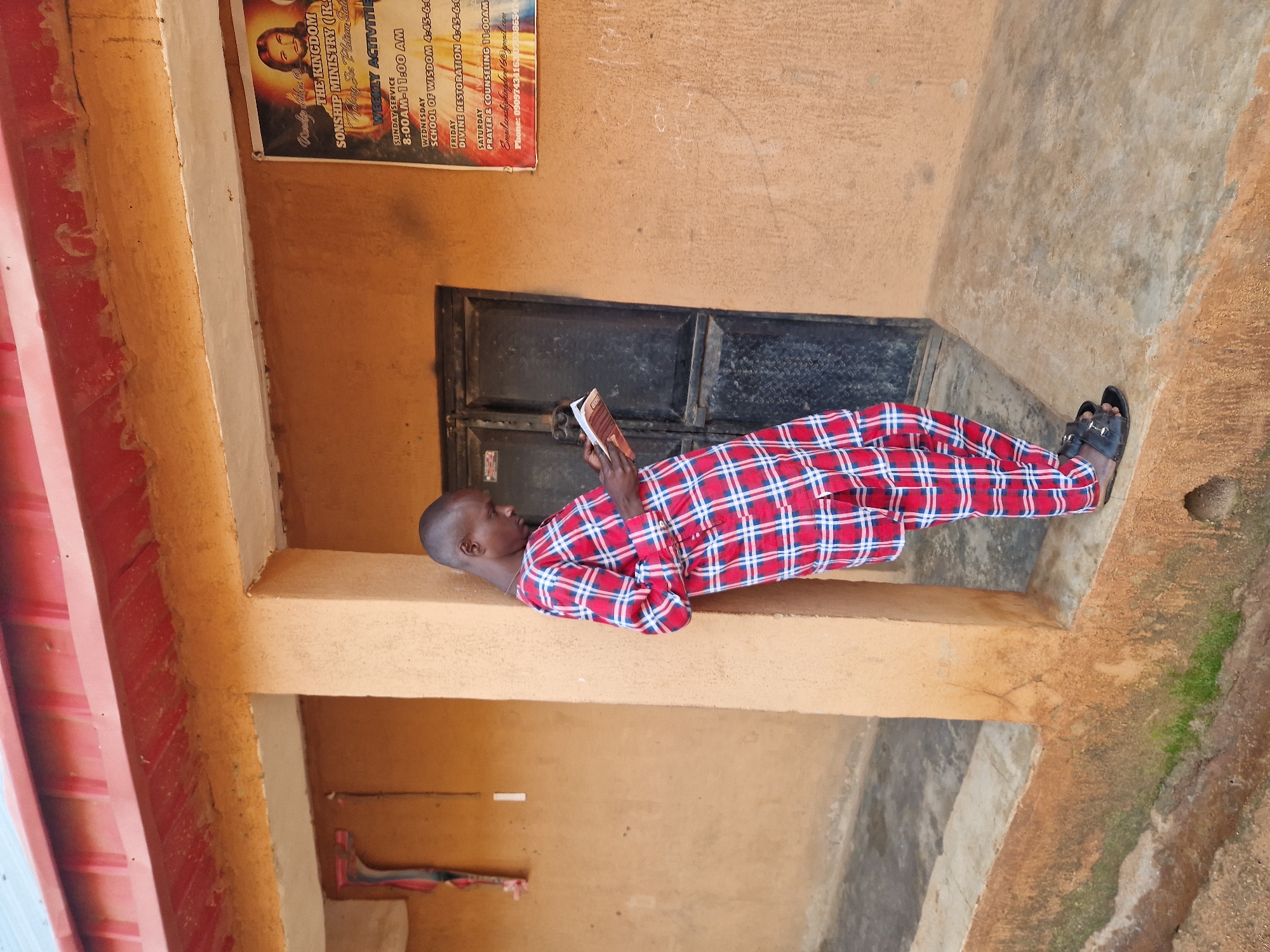 A man, identified as a resident of Jos, Nigeria, stands leaning against a textured, orange-colored building wall. He is wearing a red and blue checkered traditional attire and black sandals. He holds a book open in his hands, appearing to read it, and his gaze is directed downwards. Behind him is a dark, metal door. A poster displaying an image of Jesus Christ and church activities is affixed to the wall to his right. The ground in front of the building is a concrete walkway, with some moss and dirt visible. The building has a red metal roof and a concrete overhang. The scene appears to be an outdoor area adjacent to a religious institution.