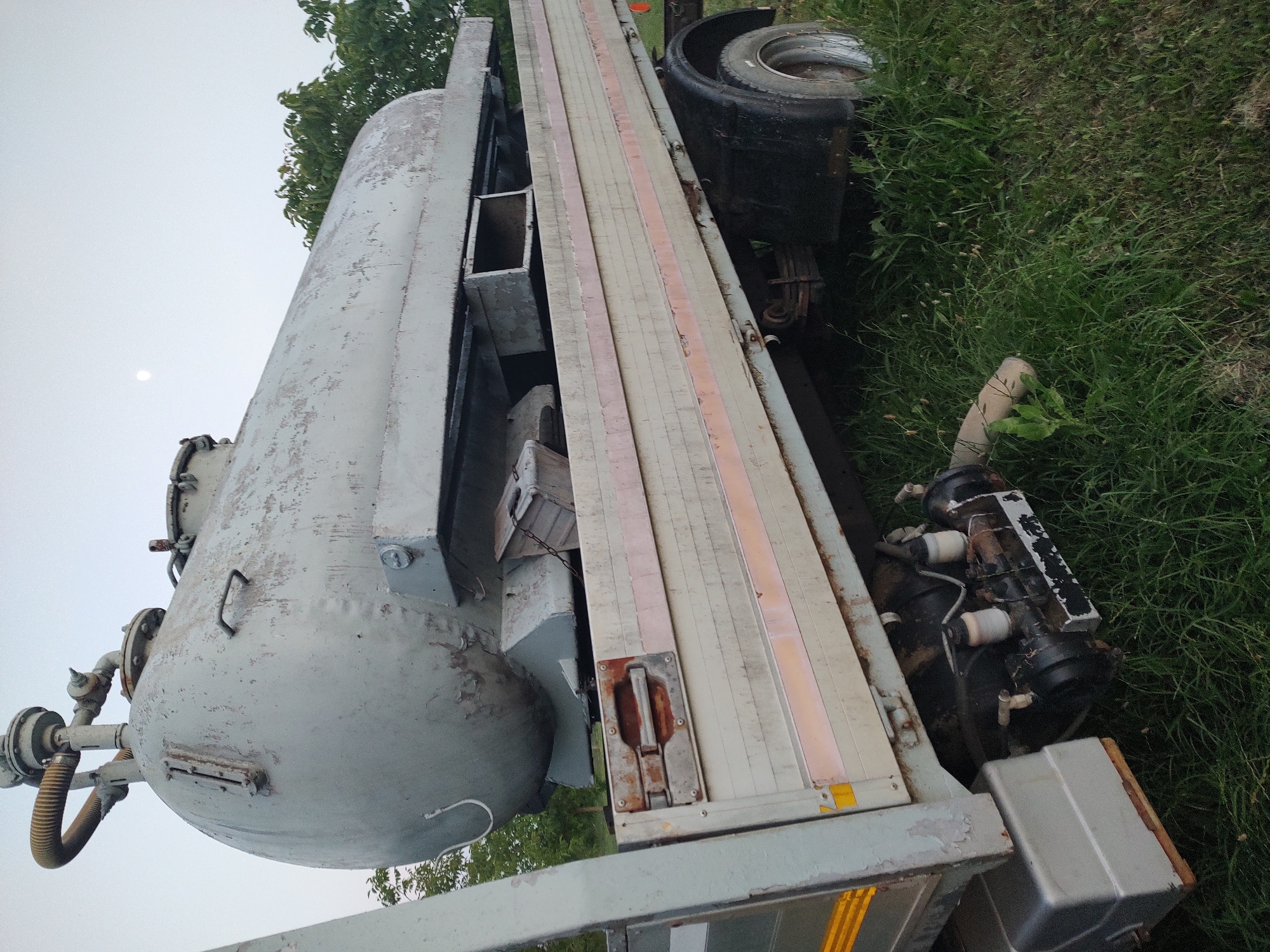 This media file captures an angled view of a large, industrial trailer with a cylindrical tank, parked on a grassy area in Kochmar, Bulgaria. The trailer appears to be made of metal and wood, with visible structural elements and a textured surface. The tank, predominantly light grey, shows signs of wear and peeling paint, suggesting it might be old or in need of maintenance.
Prominent features on the trailer include various pipes and valves attached to the tank, as well as a large tire and suspension system visible near the rear. A section of the trailer's bed is covered with wooden planks, bordered by what appear to be metal strips. The surrounding environment is characterized by lush green grass and foliage, indicating an outdoor setting. A hint of a bright sky suggests it is daytime, possibly late afternoon given the lighting.
The overall impression is one of a stationary, possibly retired or awaiting repair, piece of industrial equipment situated in a natural, unpaved location. No people are visible in the frame, and no specific activity or event is depicted. The image focuses entirely on the inanimate objects and their immediate surroundings, conveying a sense of stillness and perhaps a touch of industrial decay within a rural or semi-rural setting. There is no visible text in the image.