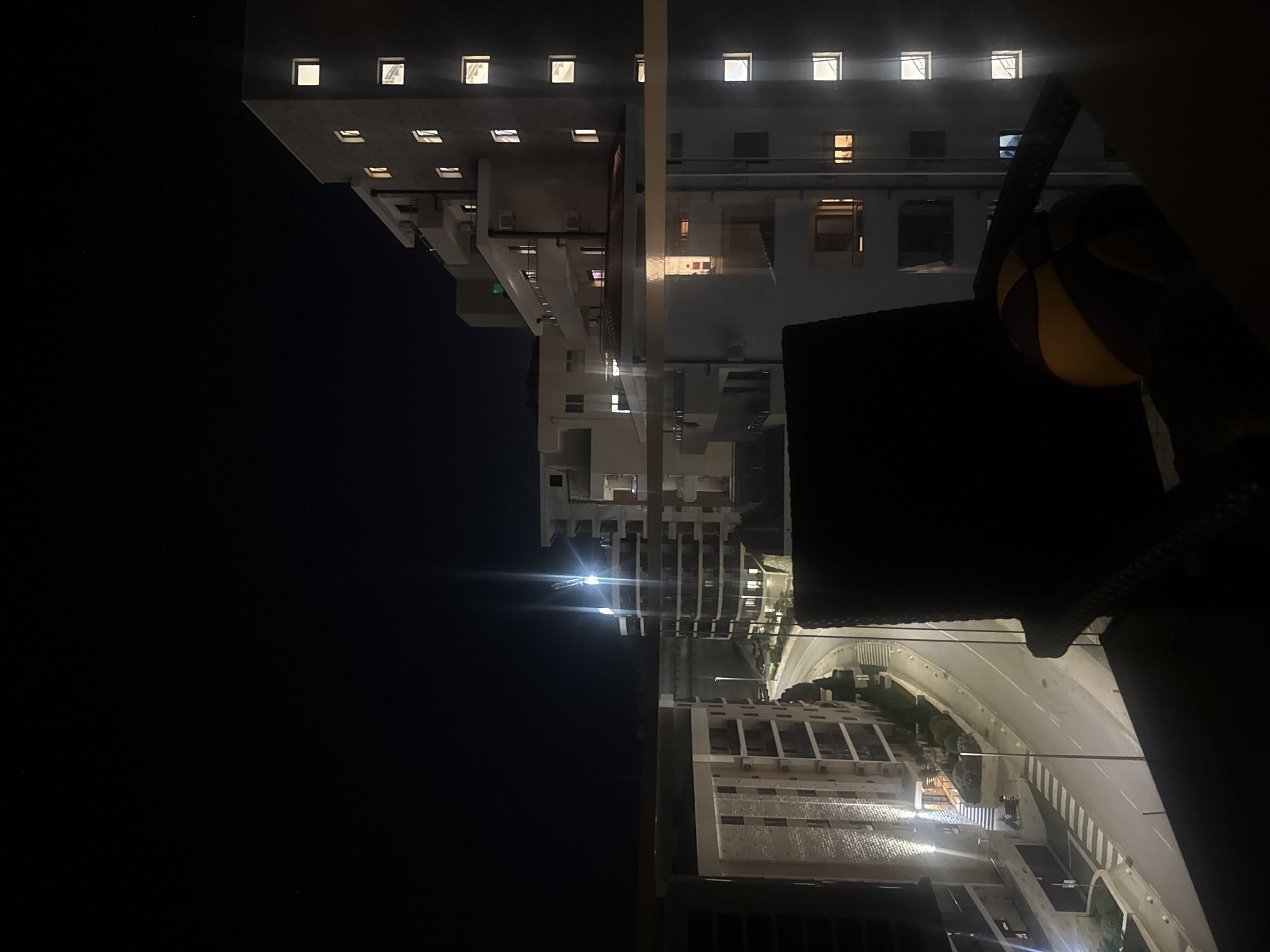 The image depicts a nighttime view of a city street in Boreti, Montenegro, from a balcony or elevated vantage point. In the foreground, a dark armchair is partially visible, with a partially obscured yellow and brown ball resting on it. A glass railing runs across the lower portion of the frame, reflecting the lights of the buildings opposite.
The scene is dominated by modern apartment buildings illuminated by artificial lights. To the left, a multi-story building with a light-colored facade features evenly spaced windows, many of which are lit. A street runs below, with a pedestrian crossing visible. Streetlights cast pools of light along the road.
To the right, another series of apartment buildings are visible, some with curved facades and balconies. These buildings also have numerous lit windows. In the background, a bright light source, possibly a construction crane or a powerful lamp, creates a lens flare effect against the dark sky. The overall impression is an urban environment at night, with the architecture and lighting characteristic of a residential area.