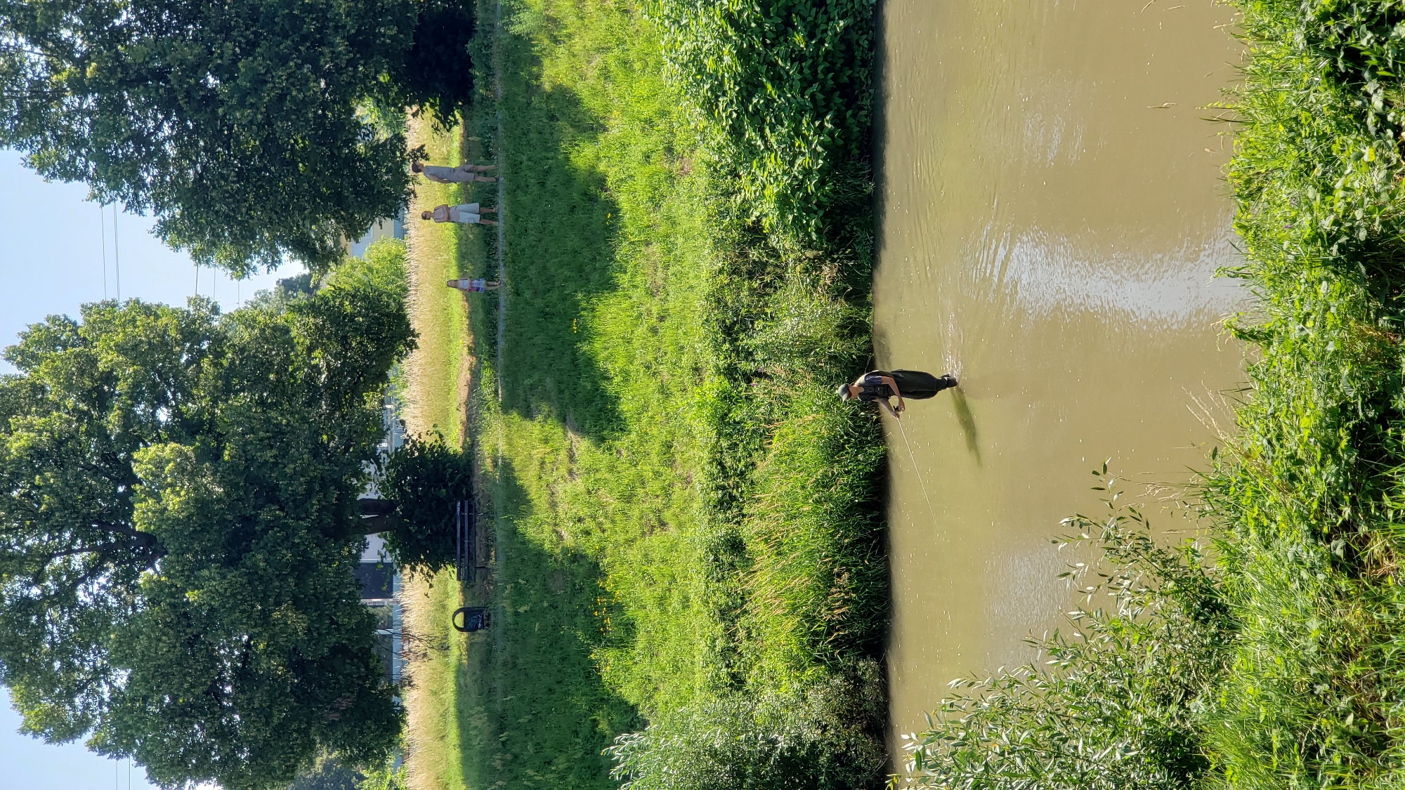 A man is fishing in a shallow, murky river in Prešov, Slovakia. He is wearing waders and a hat and is holding a fishing rod. The riverbanks are covered in lush green grass and trees. In the background, on the bank, there are a few people standing and observing. There is also a bench and a trash can under a large tree. The sky is clear and blue.