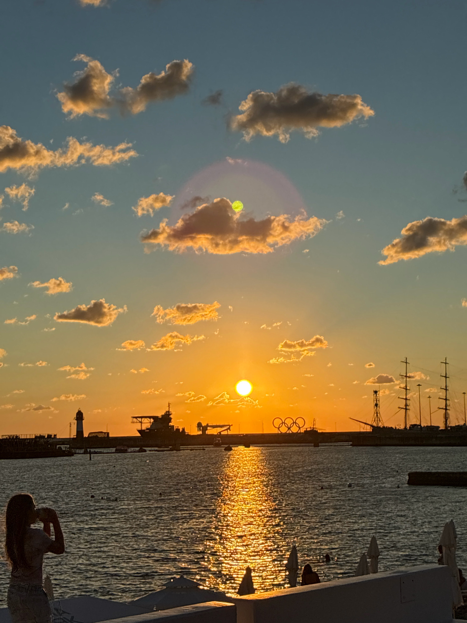 A serene sunset scene unfolds over the Black Sea in Sochi, Russia. The sun, a brilliant orb, hangs low in the sky, casting a golden glow that reflects shimmeringly on the water's surface. The horizon is dotted with various vessels, including a lighthouse, a large ship, and a tall-masted sailing ship, all silhouetted against the vibrant sky. In the distance, the iconic Olympic rings are visible, a testament to Sochi's status as a host city. A young girl stands in the foreground, silhouetted as she sips from a cup, her presence adding a touch of human element to the vast seascape. The sky is adorned with scattered clouds, tinged with hues of orange and yellow by the setting sun. The overall atmosphere is one of tranquility and natural beauty, with a subtle reminder of the city's modern legacy.