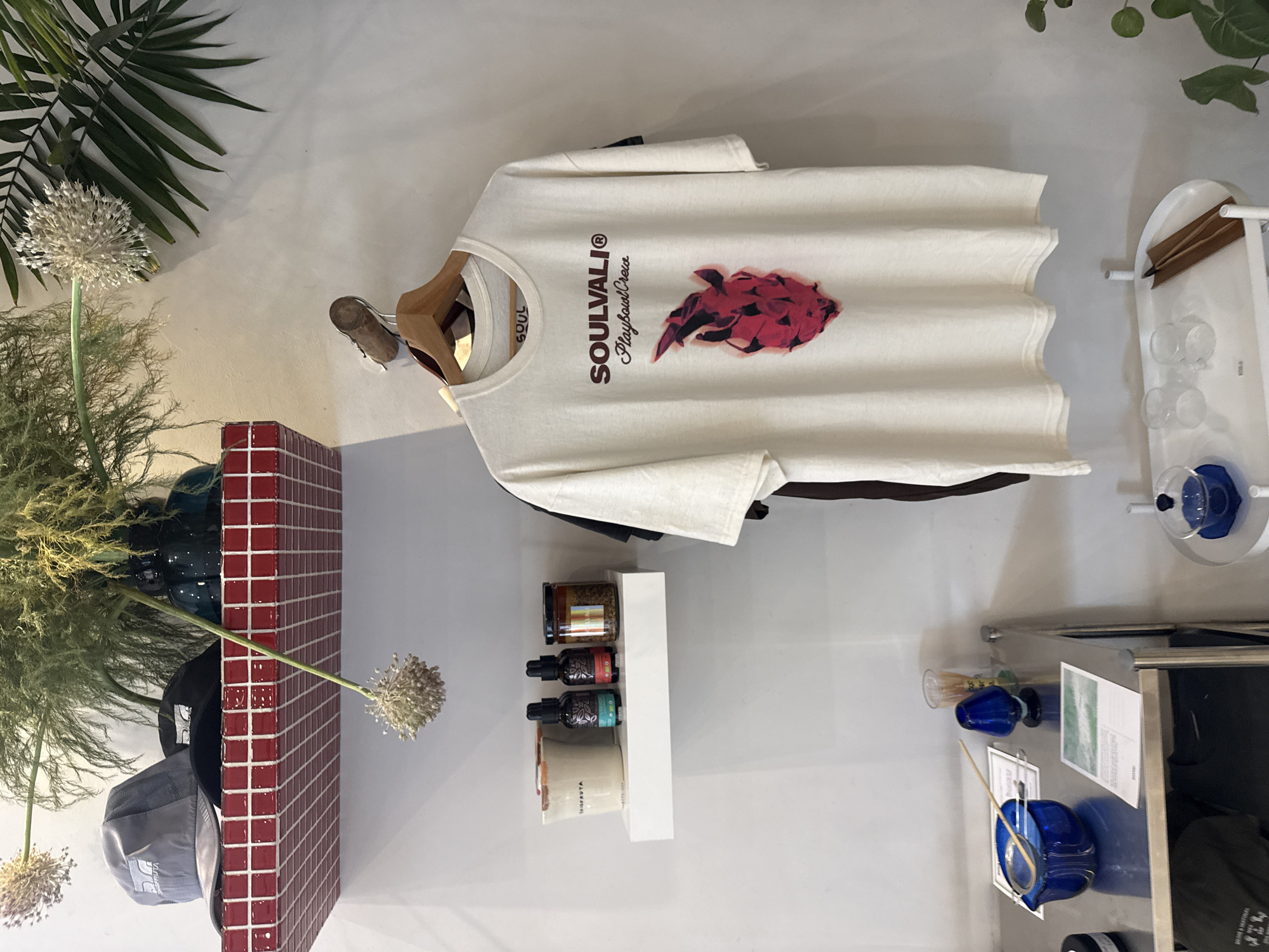 The image shows an interior scene with a white t-shirt hanging on a wooden hanger on a white wall. The t-shirt has "SOULVALI® Playsoul Crew" printed on it, along with a red abstract graphic. A red tiled structure and a plant are positioned on the left. A white shelf with jars and bottles is on the right side of the frame, with some blue glass objects and other display items also in the scene. The location is likely a retail or boutique setting, but there is no specific indication of being in the city of Puebla, Mexico.
