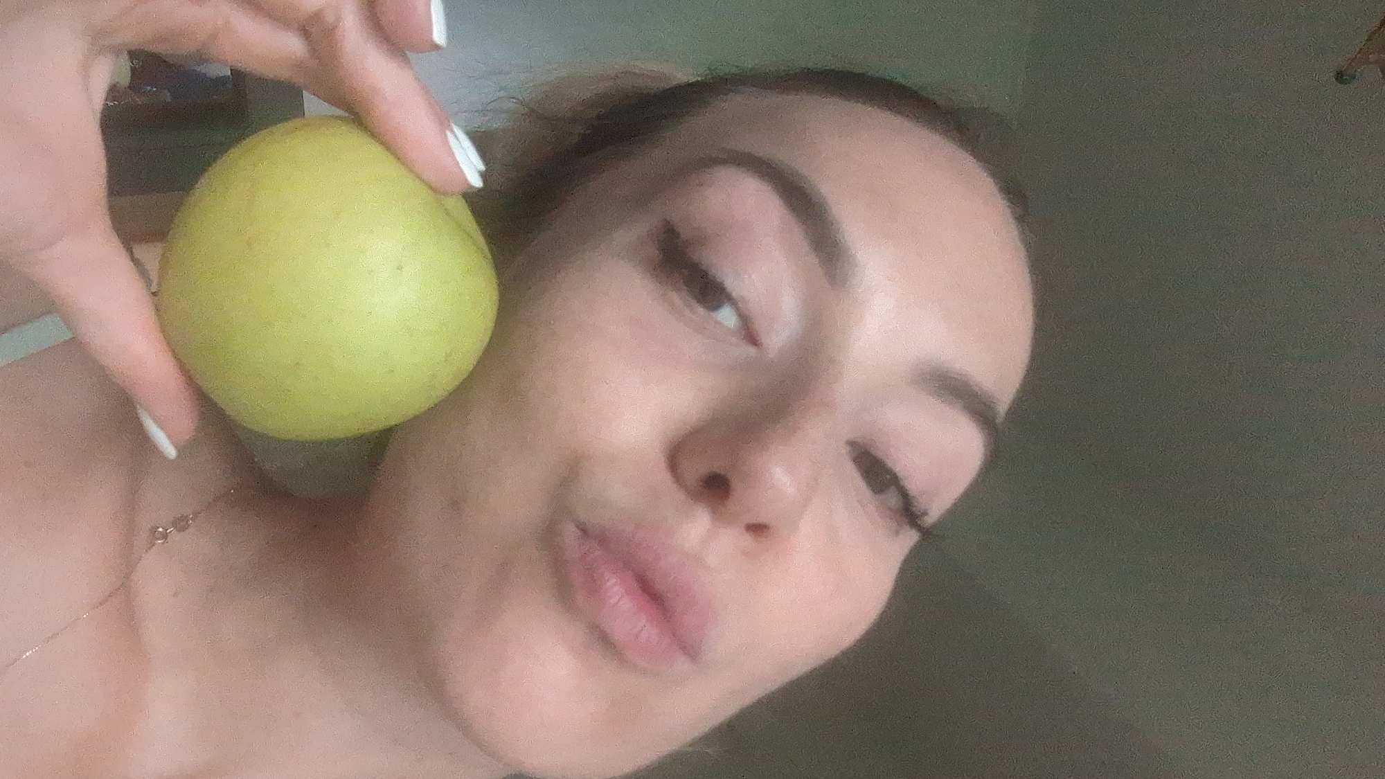 A close-up, slightly angled selfie shows a woman holding a pale green apple. The woman's face is partially visible, with her left eye looking towards the camera and her lips pursed. She has dark eyeliner and defined eyebrows. Her nails are painted white. The background is a plain, dark gray wall. No specific location in Chișinău, Moldova can be determined from the image.