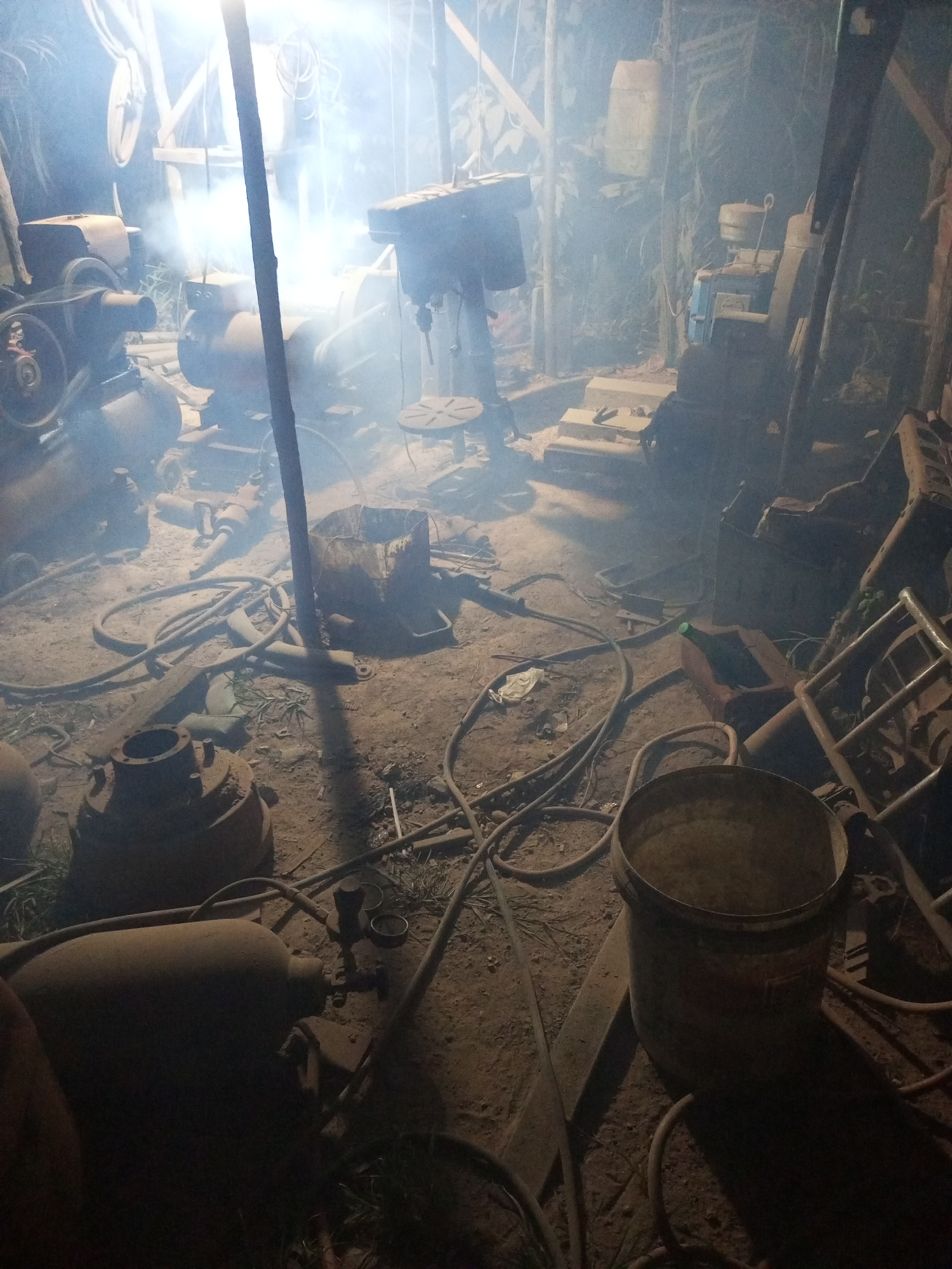 The image depicts a dimly lit, cluttered workshop in Keling Panggau, Indonesia. The scene is filled with various pieces of industrial equipment and scrap metal, including what appears to be an air compressor, a drill press, and various tanks and hoses scattered across the dusty, dirt floor. A thick haze of smoke or dust hangs in the air, illuminated by an overhead light source, creating a somewhat ethereal and obscured atmosphere. The workshop is packed with objects, suggesting ongoing work or storage of materials. The overall impression is one of a functional but untidy workspace. There are no visible people in the image.
