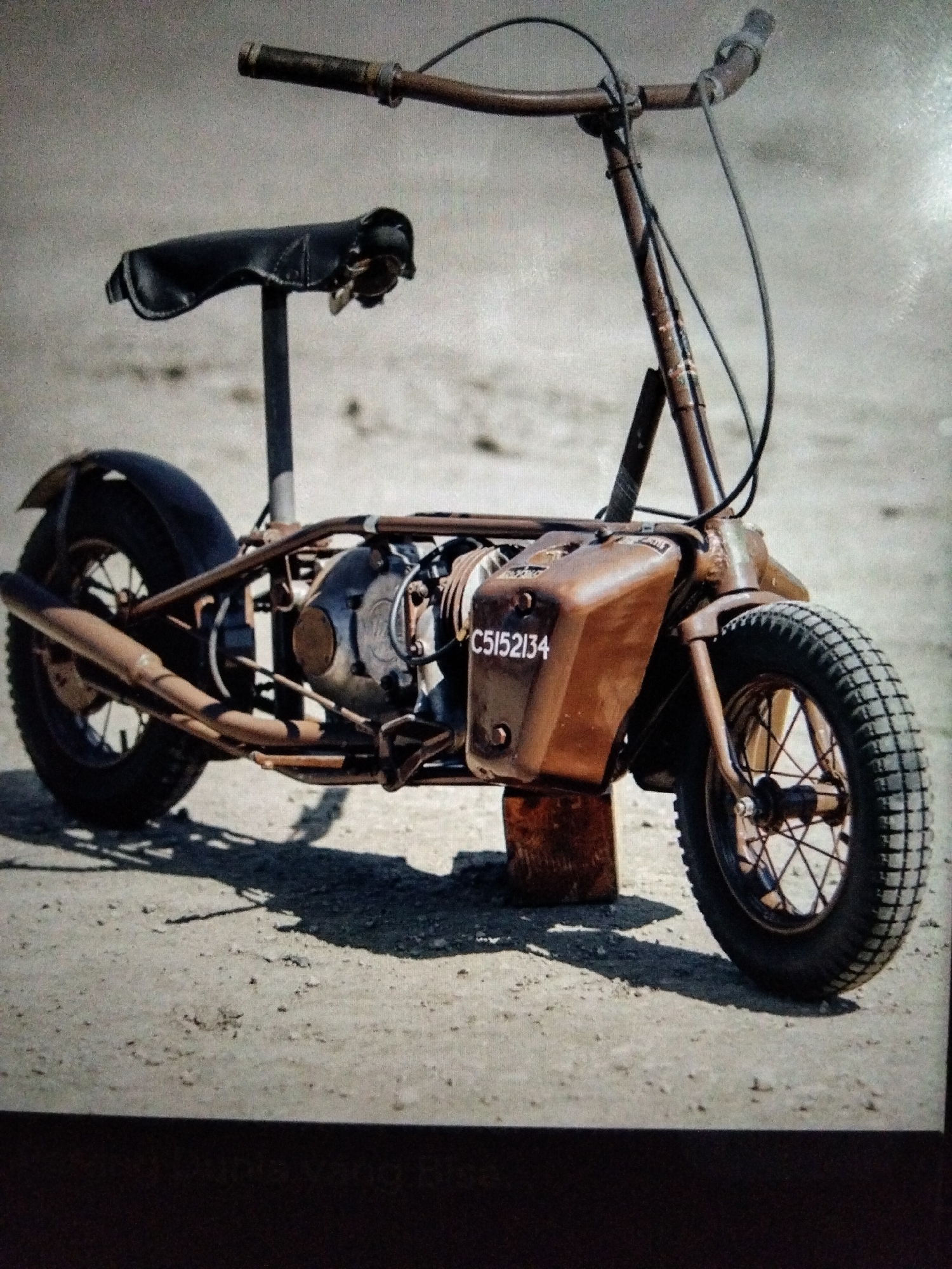 A rusty, brown motor scooter with the license plate C5152134 sits parked on a sandy surface, possibly a street in Kualaraja, Indonesia. The scooter is a vintage model with a small engine, a black leather seat, and a single exhaust pipe. The handlebars are raised and appear to be made of metal. The wheels are thick and have knobby tires. The scooter is leaning slightly to the right, supported by a wooden block. The background is blurred, but suggests an outdoor environment with sandy ground. There are no people or animals visible in the image.