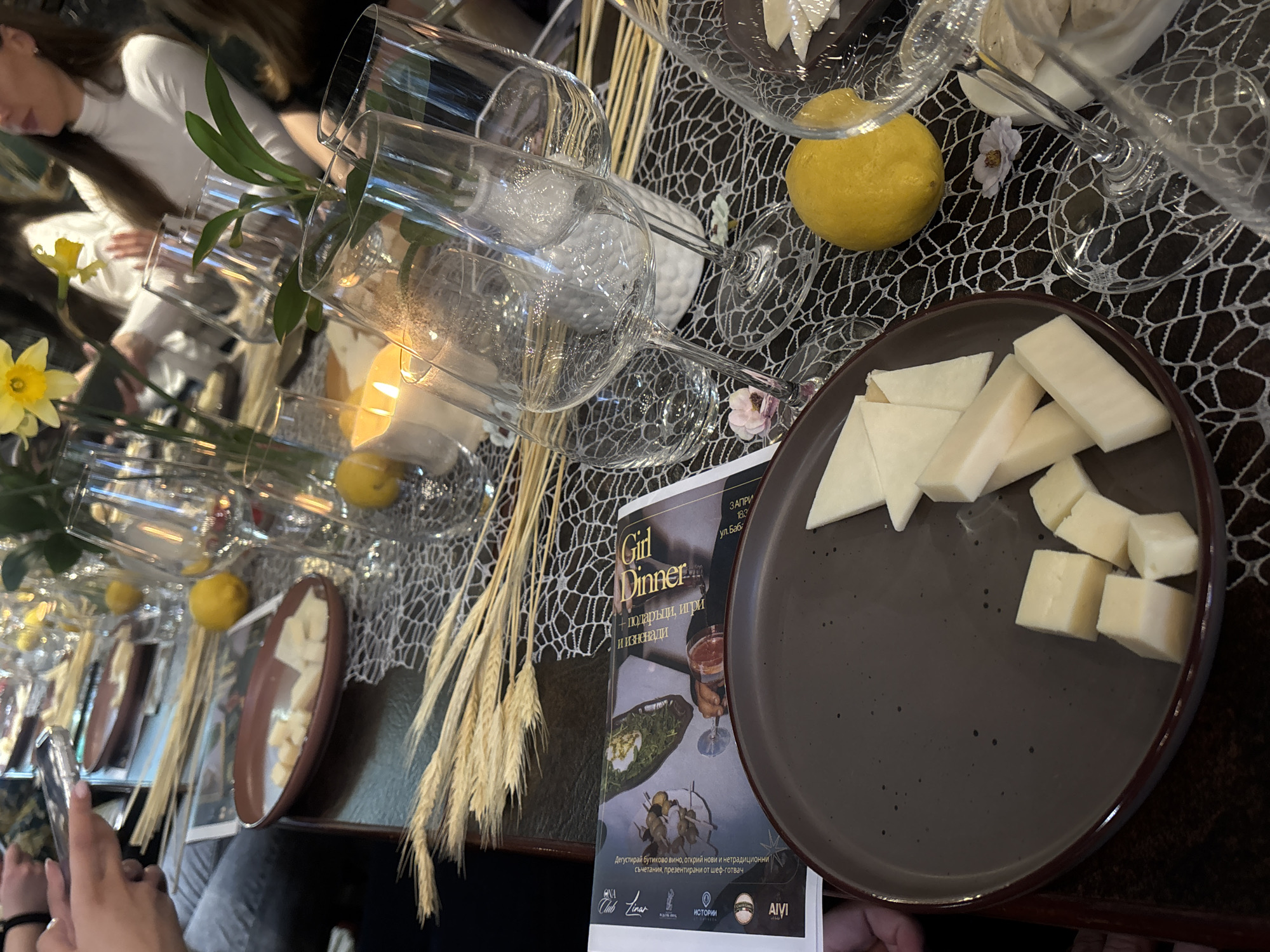 This image depicts a table setting for a "Girl Dinner" in a dimly lit environment. The table is adorned with a dark-colored, web-patterned tablecloth. Numerous wine glasses are placed on the table, along with lemons, candles, and wheat stalks as decorative elements. Plates with white cheese are also visible. A booklet or menu titled "Girl Dinner" in Bulgarian ("Girl Dinner + подаръци, игри и изненади") is present, indicating "gifts, games and surprises". The menu also mentions "Дегустирай бутиково вино, открий нови и нетрадиционни съчетания, презентирани от шеф-готвач" which translates to "Taste boutique wine, discover new and non-traditional combinations, presented by the chef". The menu indicates the location is Запри, 183 ул. Баб in an unidentified city in Bulgaria. Logos for "Linar Club", "ИСТОРИИ", and "AIVI" are visible. Hands are visible on the edges of the frame, suggesting someone is taking the photograph. People are discernible in the background, seemingly seated at the table. The image is taken from a high-angle perspective.