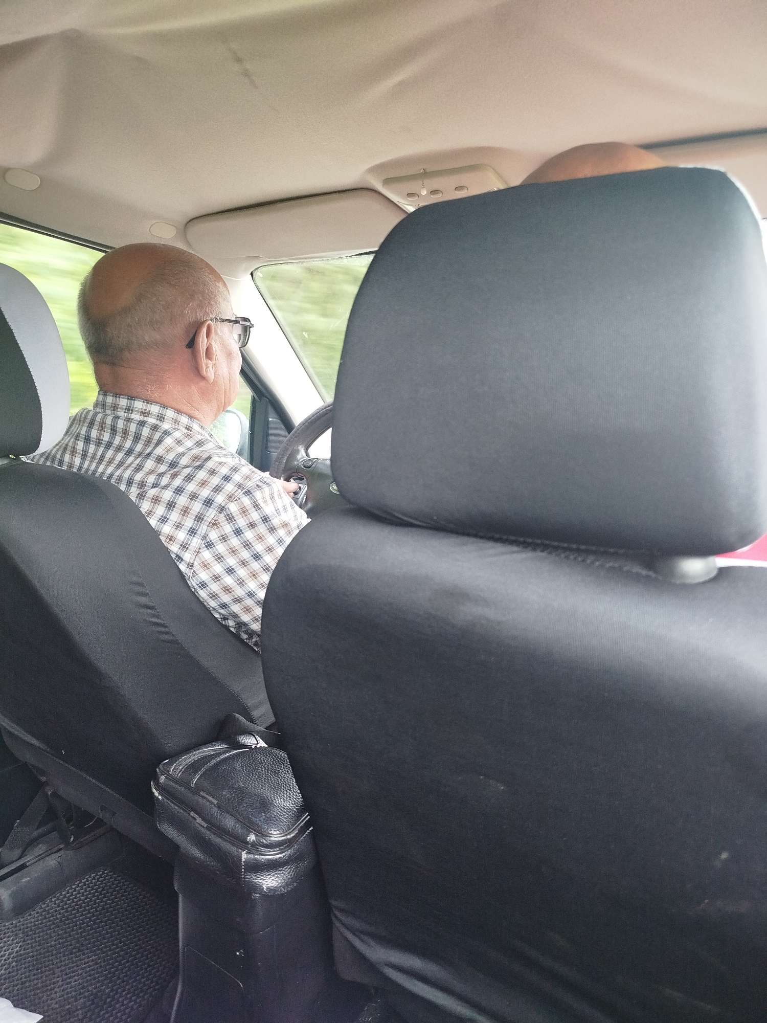 The image shows the interior of a car, as seen from the back seat. A man, appearing to be of middle age or older, is driving the car. He has a bald head, glasses, and is wearing a plaid shirt. The car's interior appears to be standard, with black seat covers. There is a leather organizer attached to the center console. The scene suggests a typical car journey, with no unusual or suspicious elements present. The location within the city of Zavodoukovsk, Russia, is not evident from the visual information in the image.