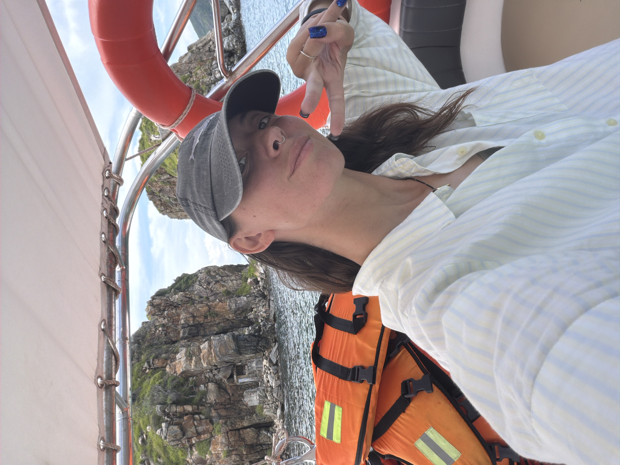 A woman wearing a striped shirt and a baseball cap is on a boat, making a peace sign with her fingers. She is also wearing a life jacket. In the background, there is a rocky cliff and water. The location is likely off the coast of Bac Nha Trang Ward, Vietnam. A life preserver is mounted near the edge of the boat.