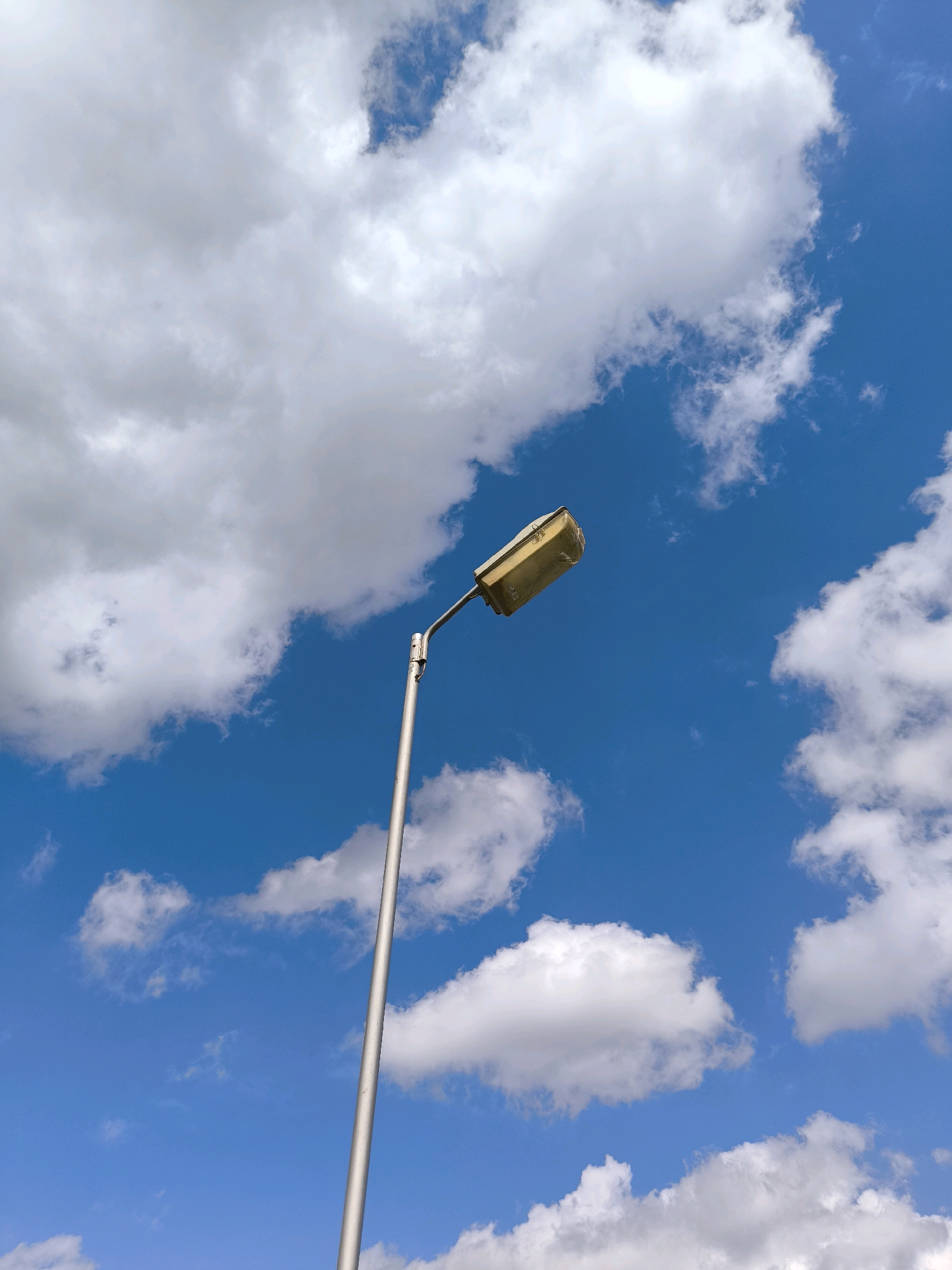 A single, silver lamppost stands against a bright blue sky filled with fluffy white clouds. The lamppost is positioned diagonally, extending from the bottom left corner towards the upper center of the frame. Atop the lamppost is a rectangular, gold-colored lamp fixture. The scene is photographed from a low angle, looking upwards. The environment appears to be outdoors, likely in a public area such as a park or street within Karanganyar, Indonesia, given the context. There are no people or animals visible in the image.