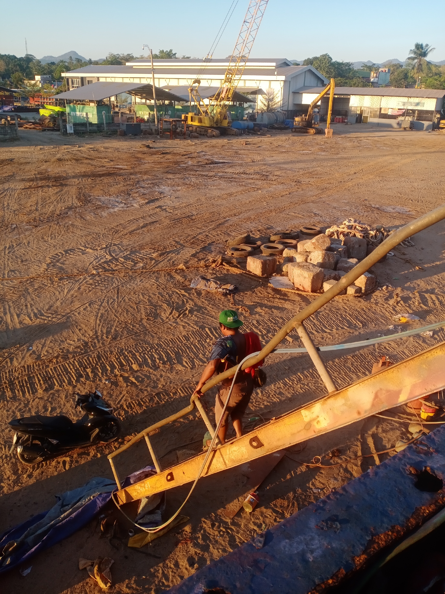 An outdoor, construction site scene in Siddo, Indonesia, shows a dirt lot with tire tracks from vehicles and heavy machinery. In the background, large buildings with metal roofs are visible, along with a yellow crane and various pieces of construction equipment. In the foreground, a person wearing a green cap, a backpack, and shorts is walking up a metal staircase. To the left of the staircase, a black motorcycle is parked. The ground is covered in dirt, sand, and scattered debris, including tires, concrete blocks, and ropes. The lighting suggests it is either early morning or late afternoon, with long shadows cast across the scene.