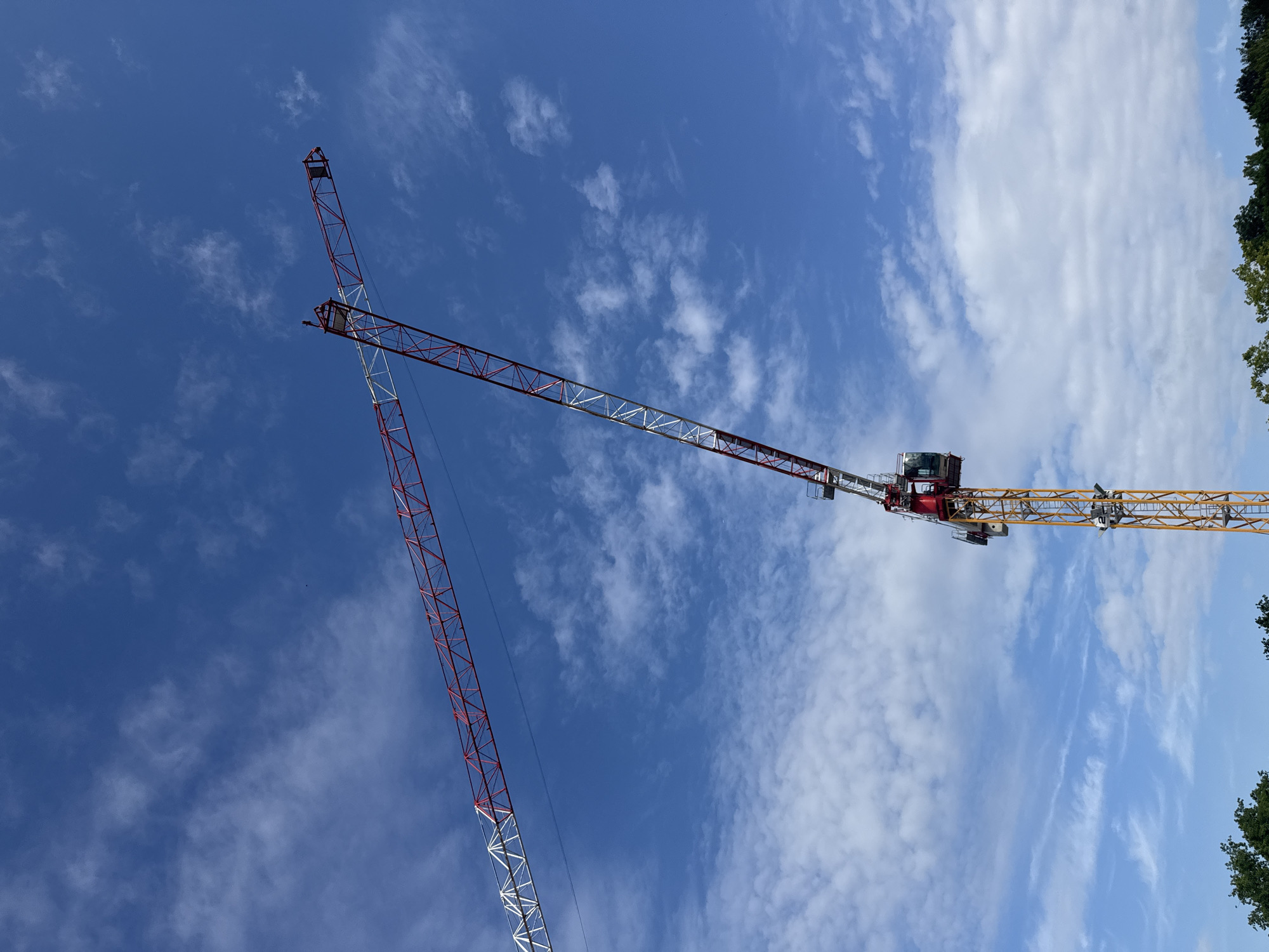 The image shows two construction cranes silhouetted against a bright blue sky with scattered white clouds. The cranes are made of a lattice structure, with one primarily red and white, and the other yellow with red accents. Both cranes are in a state of construction or deconstruction, with one crane's boom angled upwards and the other's boom extending across the frame. The perspective is from below, looking up at the cranes and the sky. A portion of green foliage from trees is visible on the right side of the image, suggesting an outdoor setting, possibly a construction site in a vegetated area. The lighting suggests it is daytime, likely late morning or afternoon, given the brightness of the sky. There are no people visible in the image. No text is present.