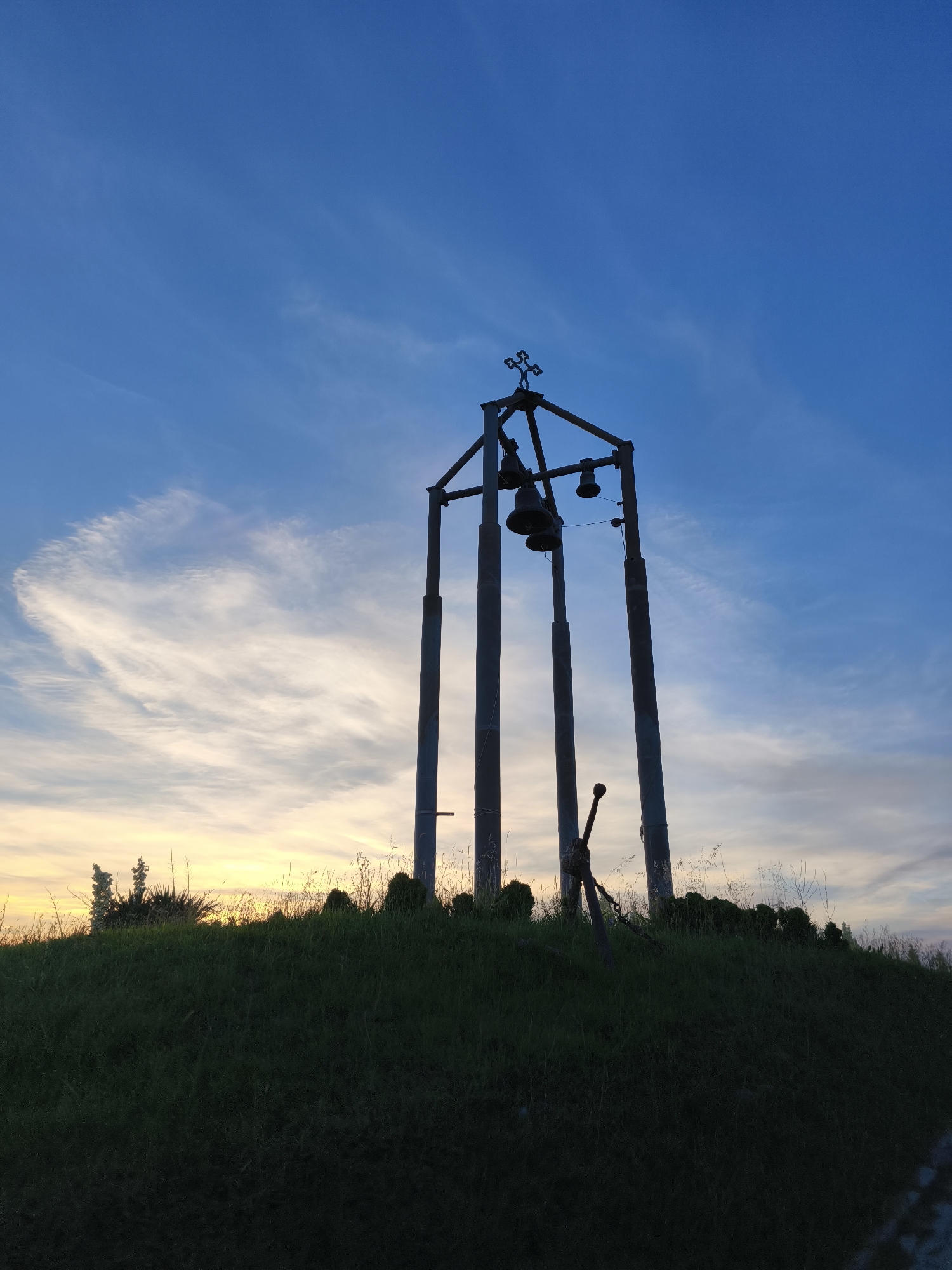 The image shows a bell tower silhouetted against a twilight sky at the highest point of a grassy hill. The bell tower is a metal structure with four vertical supports and a cross at its apex. Three bells are suspended from the crossbar of the tower. A mechanism, possibly for ringing the bells, is visible attached to one of the supports. The hill is covered in green grass and sparse vegetation, with the faint outline of some trees or bushes at the crest. The sky is a gradient of deep blue at the top, transitioning to lighter blue and then to yellow and orange hues near the horizon, indicating sunset or sunrise. Wispy clouds are scattered across the sky. The scene appears to be located on the outskirts of Tsarevo, Bulgaria, likely a public space or a landmark. No people are present in the image.