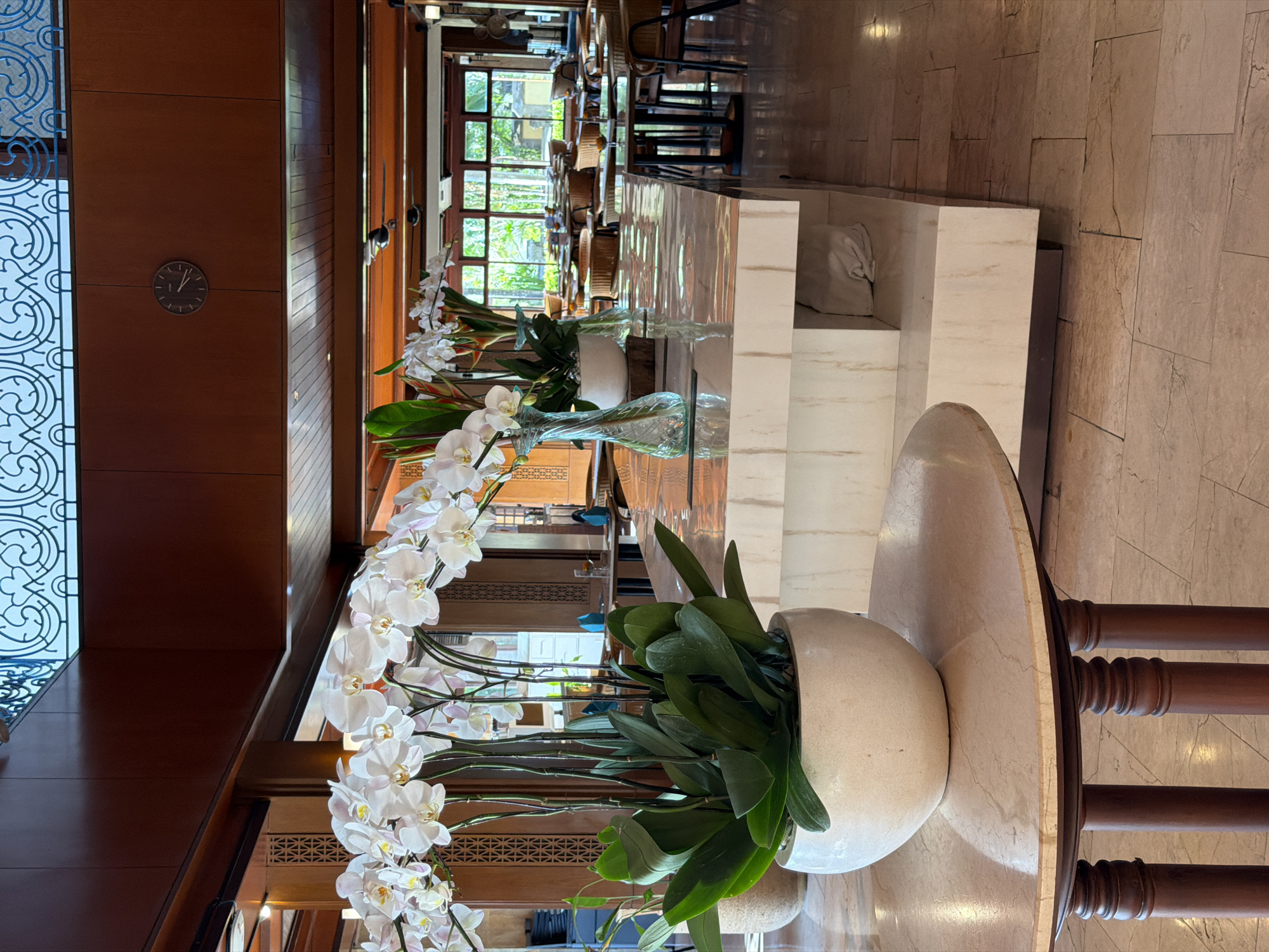 The image shows a restaurant interior, possibly in Kuta, Indonesia. The foreground features a round marble table adorned with a white pot containing orchids and green plants. Behind the table is a marble bar counter with glass vases of white flowers. The background shows a well-lit dining area with wooden furniture, and large windows offer a view of a tropical environment. On the left, a wooden wall is visible with a decorative clock and ornate design details. The restaurant's decor suggests an upscale ambiance.