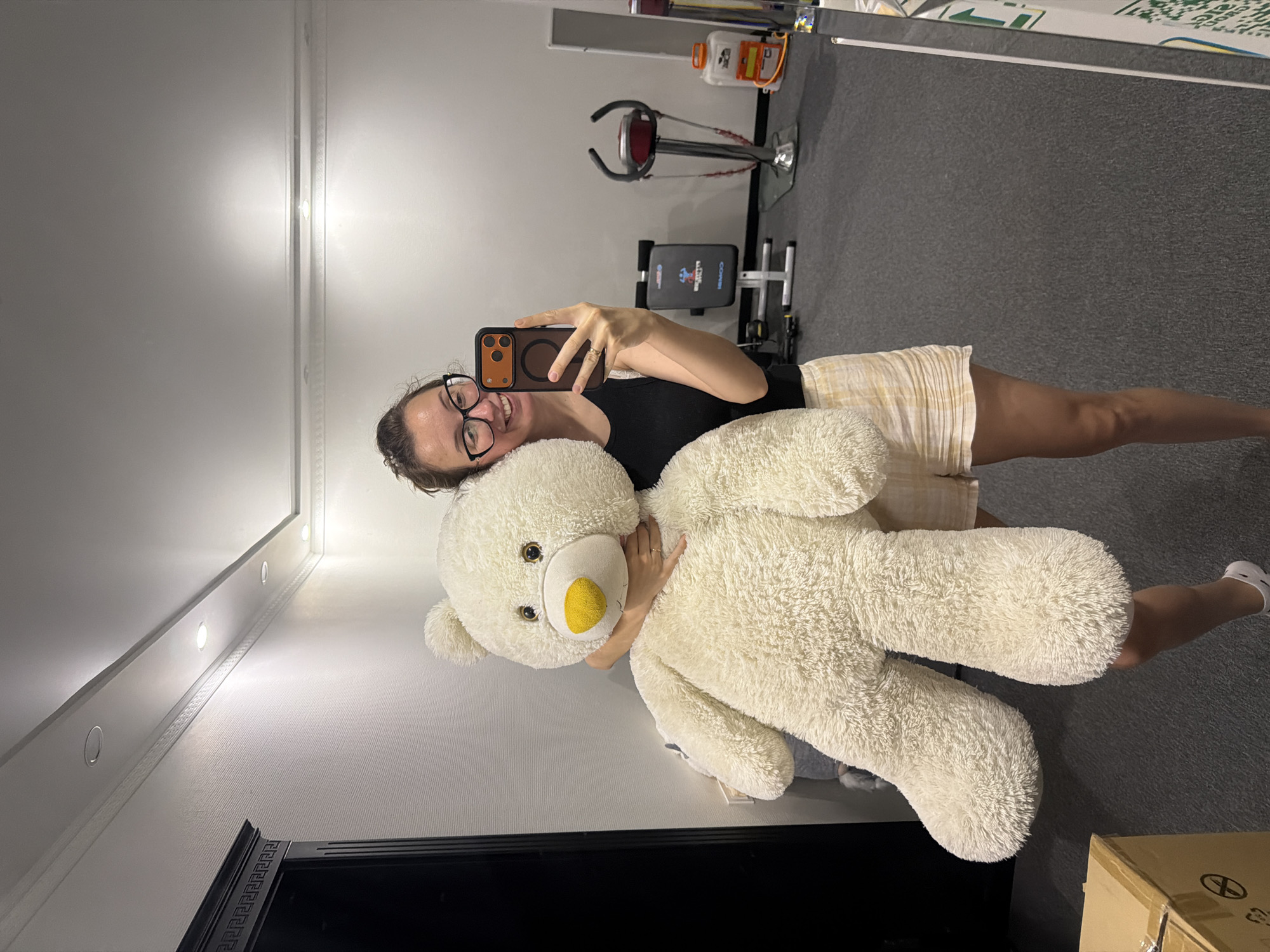 A woman stands in what appears to be a home gym, holding a large, fluffy, cream-colored teddy bear. She is wearing a black tank top and plaid shorts, and glasses. She is taking a selfie with her phone, which has an orange case. In the background, there is a black wardrobe, a weight bench, and a vibrating platform. There is also a box in the lower left corner of the image. The lighting suggests it is daytime, but the environment is indoors.