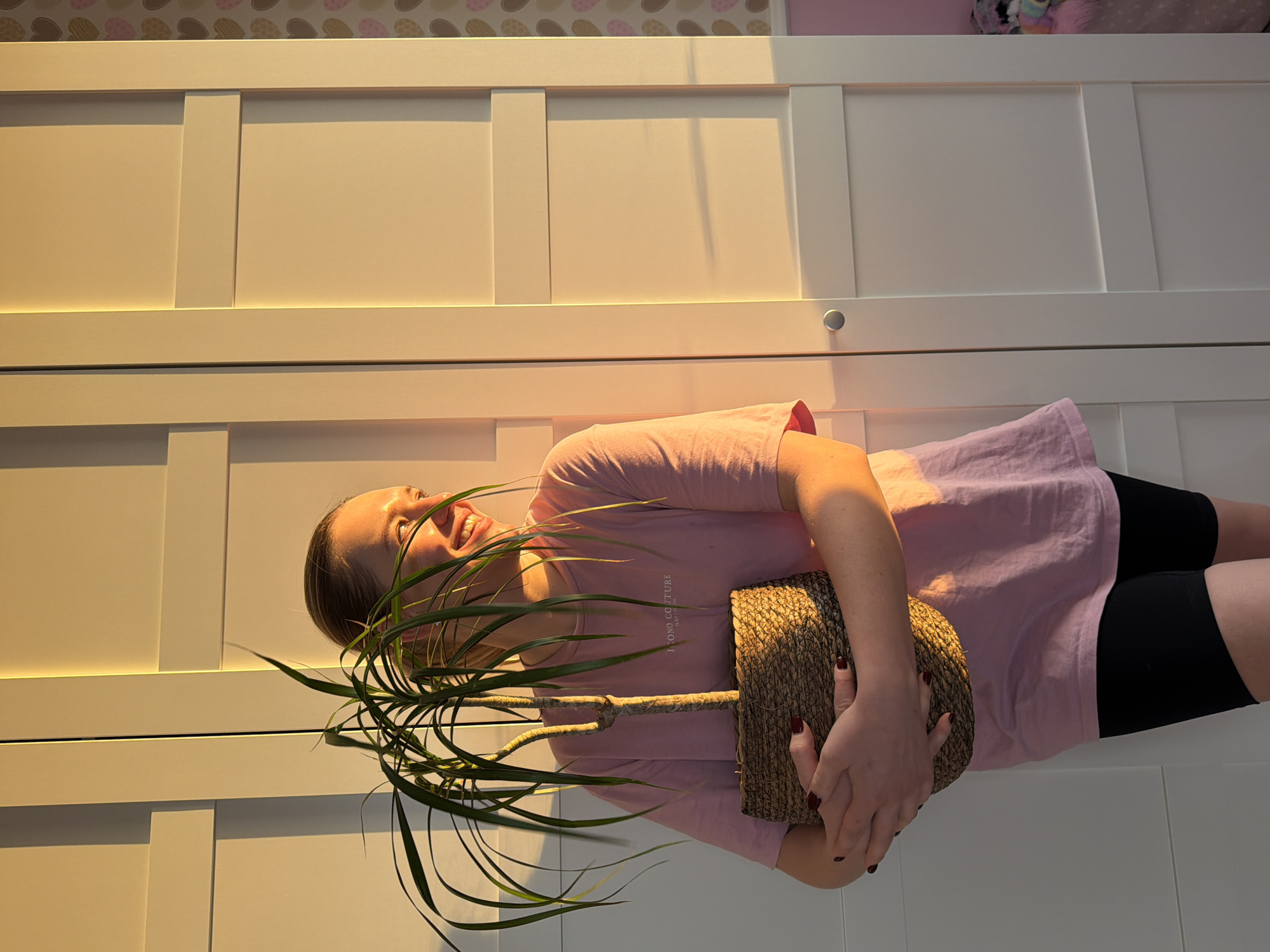 A woman in a pink t-shirt and black shorts is holding a potted plant in front of a white wardrobe. The wardrobe has a panelled design. A wallpaper with repeating heart shapes is visible above the wardrobe. The image appears to be taken indoors, possibly in a residential setting in Dreiliņi, Latvia, though no specific landmarks confirm the city directly.