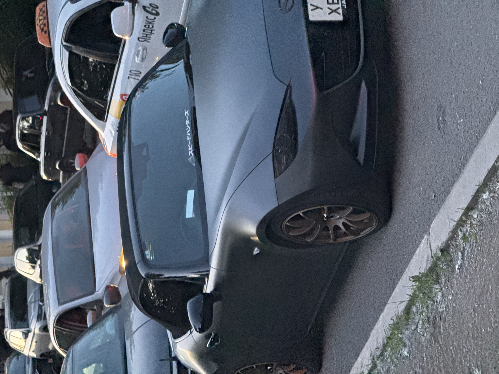 The image shows a street scene in Yekaterinburg, Russia, with several cars parked and in motion. In the foreground, a dark gray Mazda MX-5 Miata convertible is prominently featured, with its top down. The car has bronze-colored wheels and a sticker with Japanese characters on the windshield. To the right of the Miata, a white Yandex Go taxi is visible, with the number "710" on its side. Behind the taxi and the Miata, other vehicles, including a silver sedan and a dark-colored car, are present. The street appears to be asphalt, with a concrete curb and some sparse vegetation along the side. The lighting suggests it is daytime.