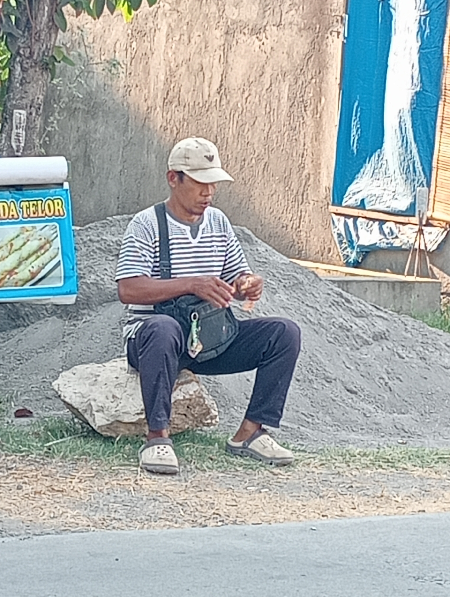 A man wearing a striped t-shirt, dark pants, and a light-colored baseball cap is sitting on a large rock next to a pile of gravel. He is holding a small object in his hands, possibly food. A dark green bag is slung across his chest. To his left is a blue sign with text and an image of what appears to be food items. Behind him is a rough, textured wall and some blue tarp material. The scene appears to be outdoors in a semi-urban or roadside environment in Tanjunganyar, Indonesia.