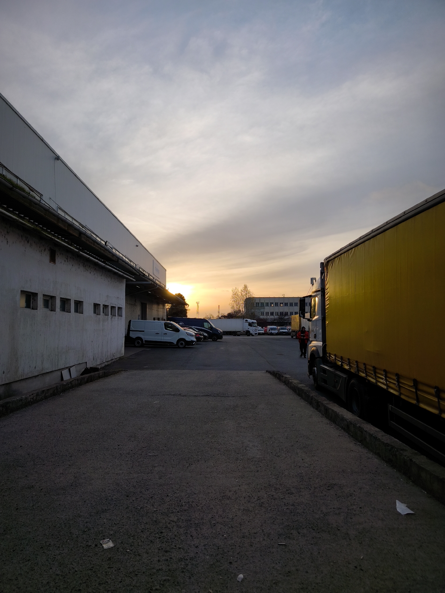 The image captures an industrial or commercial yard in Burgas, Bulgaria, during either sunrise or sunset, indicated by the warm, golden light on the horizon contrasting with the shadowed foreground. The sky above transitions from a light blue-grey with scattered clouds to vibrant orange and yellow near the sun.
On the left, a large building with a corrugated grey facade stands, featuring a lower concrete section with a row of small, rectangular windows. Pipes and drainage systems are visible along its upper edge. The ground in the foreground and mid-ground is a paved asphalt surface, showing some wear, with curbs lining the edges. A small piece of white litter is visible on the ground in the bottom center.
Various vehicles are parked throughout the area. A prominent yellow semi-trailer truck, with a bright yellow side tarpaulin, occupies the right side of the frame, its cabin partially visible. Further back and to the left, several vans, including a white one and darker models, are parked in a line. In the distance, another white semi-trailer truck is faintly visible.
A person wearing a red or orange jacket walks across the mid-ground, partially obscured by the yellow truck, adding a human element to the otherwise industrial landscape. Another figure can be vaguely seen near the distant white truck. Bare trees are visible in the background, suggesting a colder season. The scene conveys a sense of quiet activity or transition within a logistics or warehouse environment. No legible text is visible.